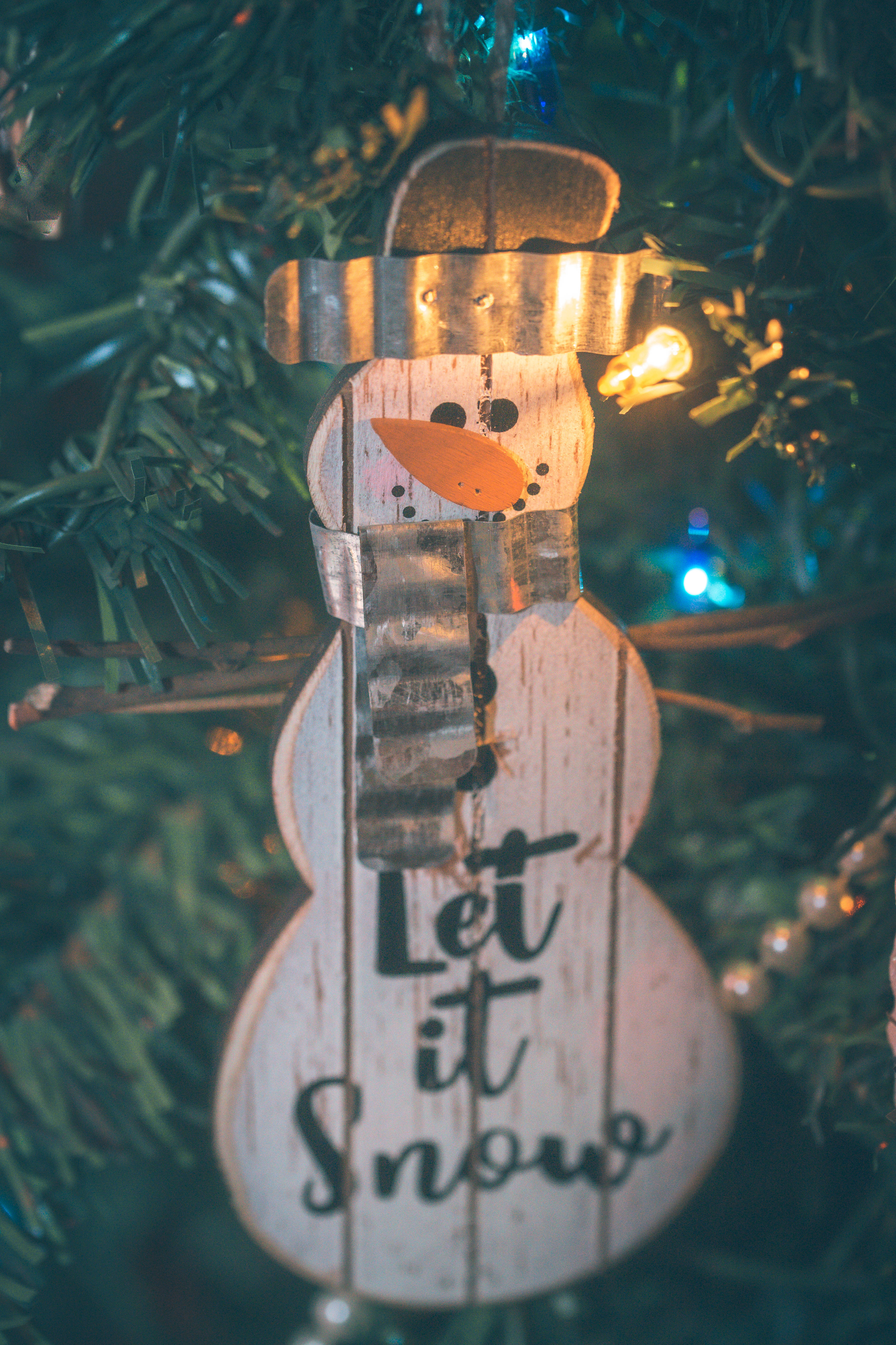 雪人 桌布 壁紙 — 一個質樸的木製雪人裝飾品，上面寫著「Let it Snow」，掛在溫暖仙女燈照亮的綠色聖誕樹上。