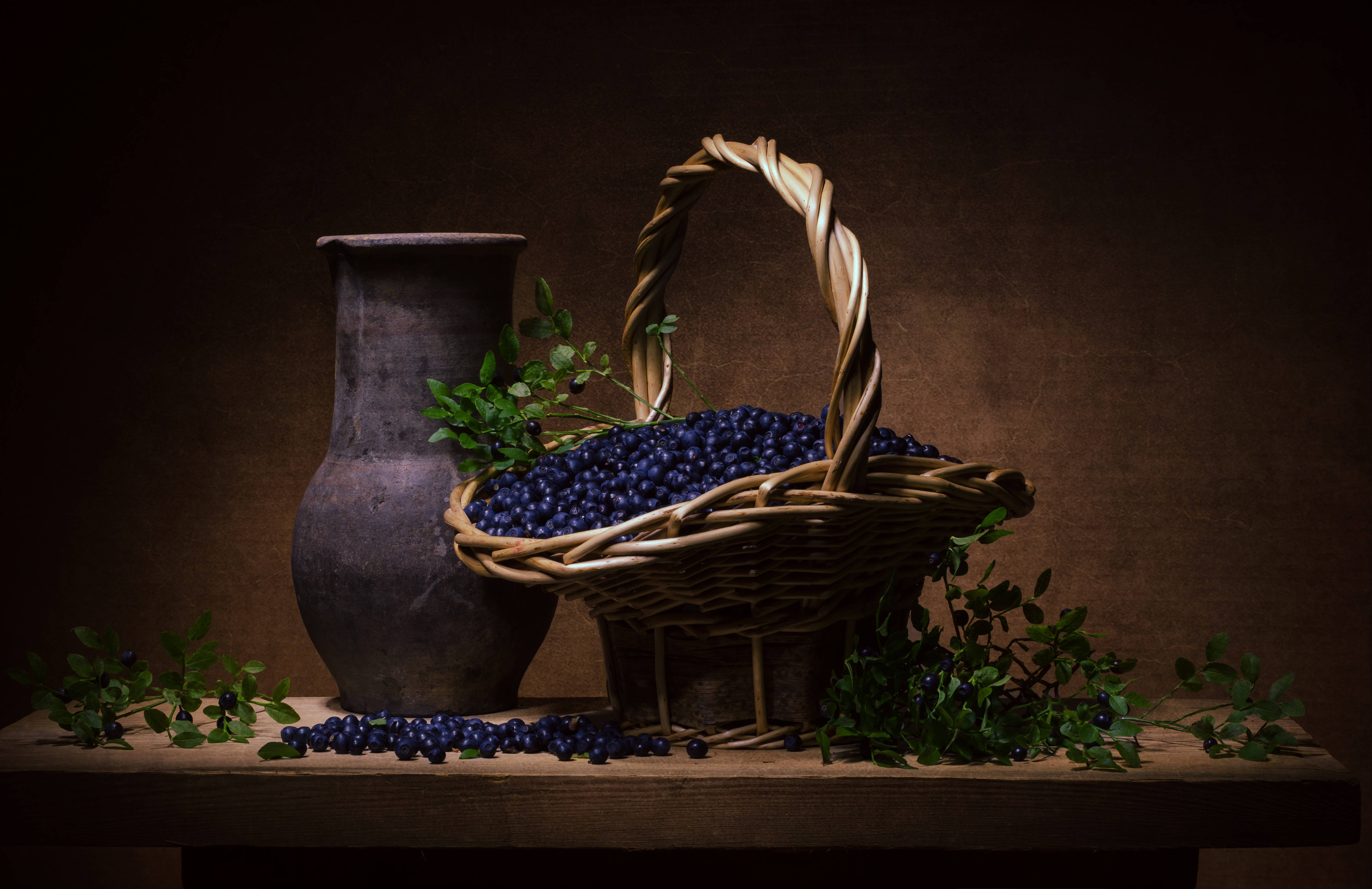 Blueberry 桌布 壁紙 — 一個質樸的柳條籃子裝滿了飽滿的藍莓，旁邊放著一個風化的陶壺在木桌上，周圍環繞著綠色藤蔓和散落的漿果，在溫暖而戲劇性的燈光下。