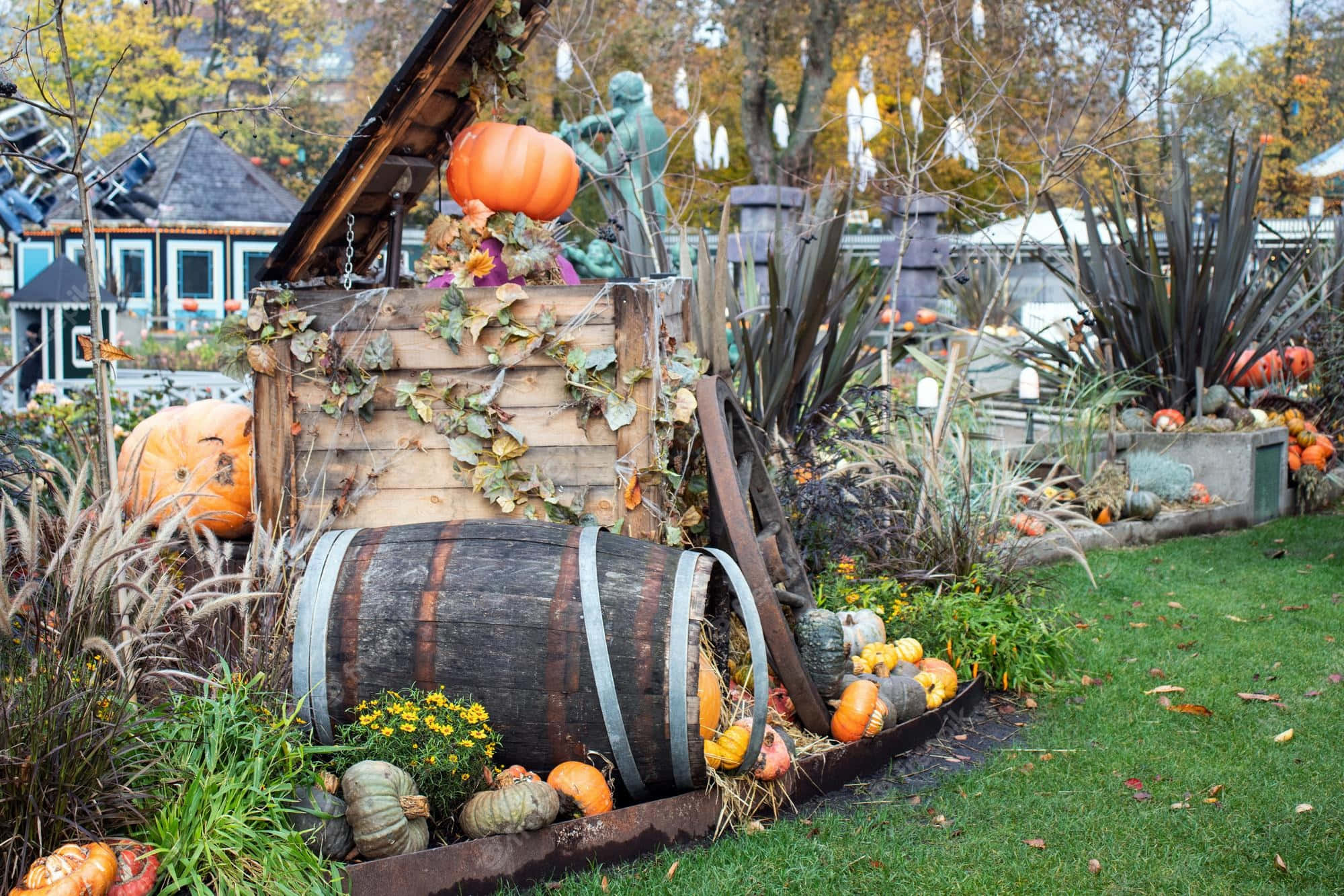 Halloween Yard Decorations 桌布 壁紙 — 一個質樸的秋季花園展示，南瓜、葫蘆和一個風化的木桶圍繞著一個帶有綠色雕像的木結構排列。