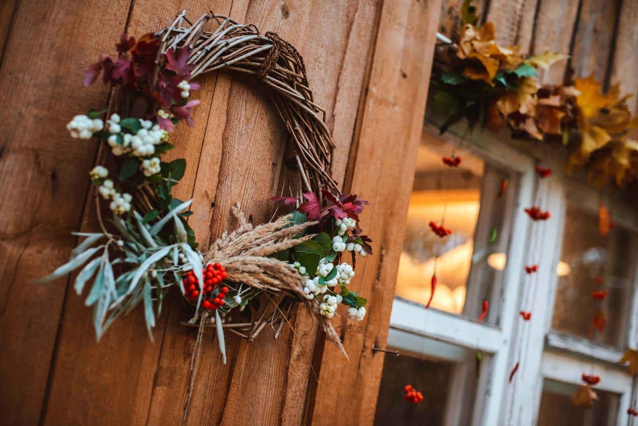 Fall Wreath 桌布 壁紙 — 一個質樸的秋季花環，帶有紅色漿果、白色漿果和乾燥樹葉，掛在風化的木門上，旁邊是裝飾著落葉的窗戶。