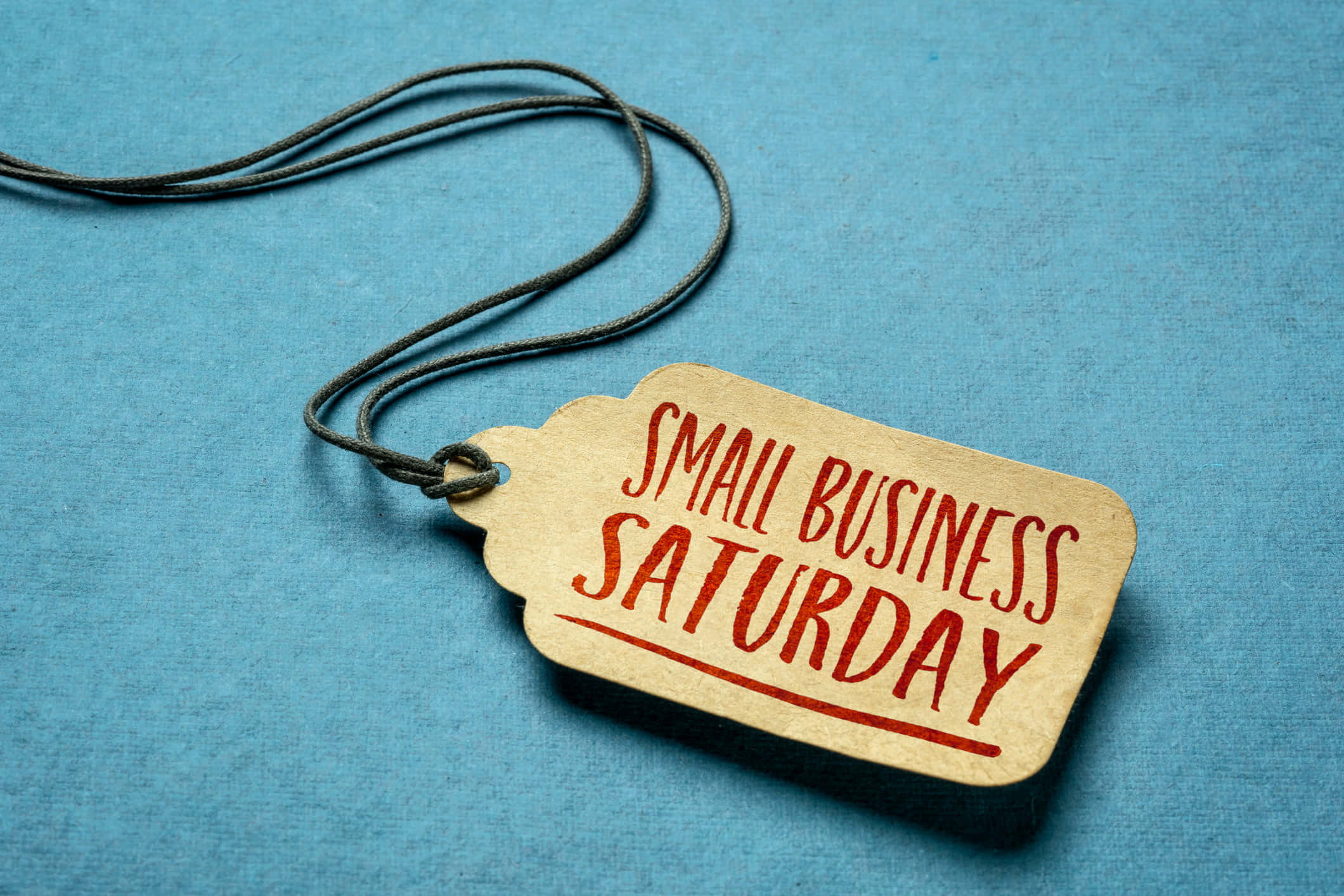 商業 桌布 壁紙 — 一個質樸的米色標籤，上面有紅色的「SMALL BUSINESS SATURDAY」文字，掛在一條深色繩子上，背景是帶紋理的藍色。
