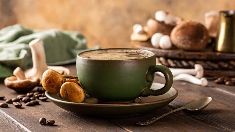 Brown Coffee 桌布 壁紙 — 一個質樸的綠色陶瓷咖啡杯放在木桌上，周圍有蘑菇、咖啡豆和復古湯匙。