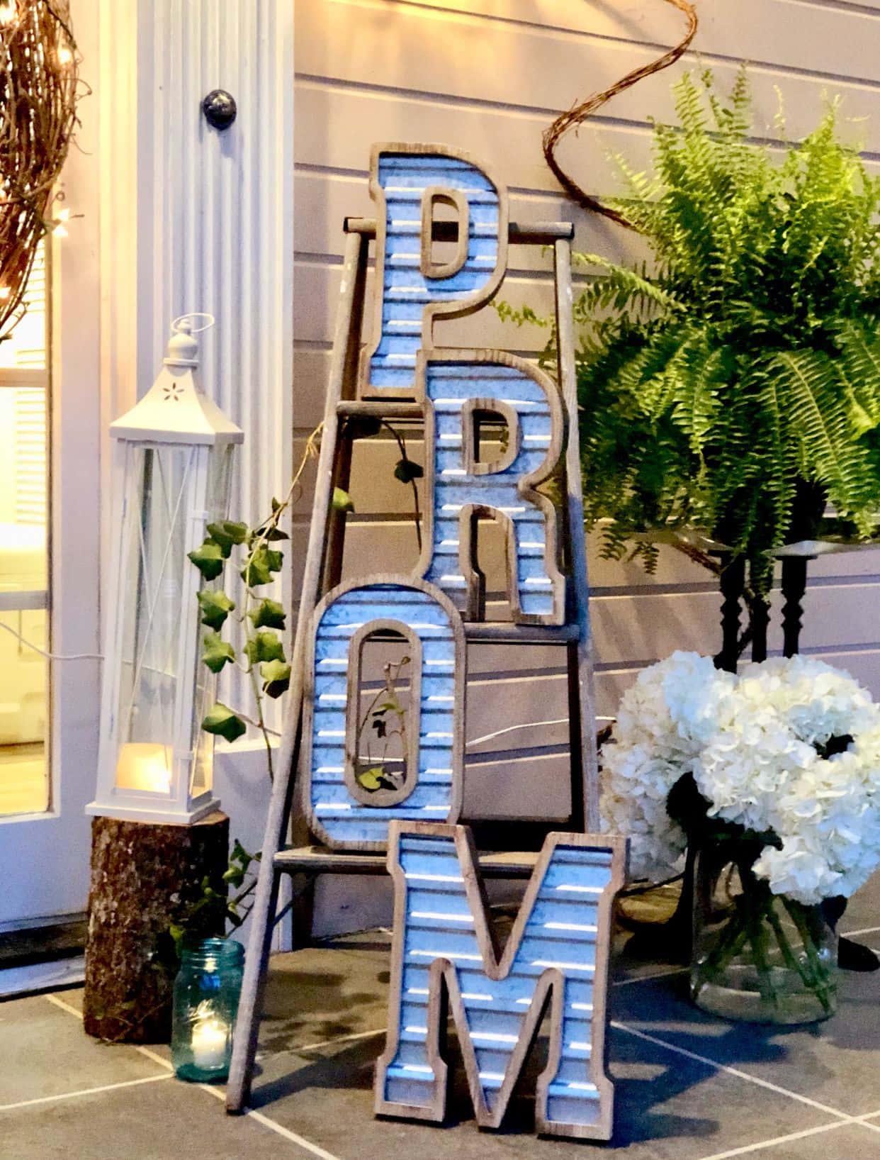 Party 桌布 壁紙 — 一個質樸的藍色「PROM」標誌矗立在戶外，旁邊有蕨類植物、白色繡球花和一個燈籠，位於石頭庭院上。