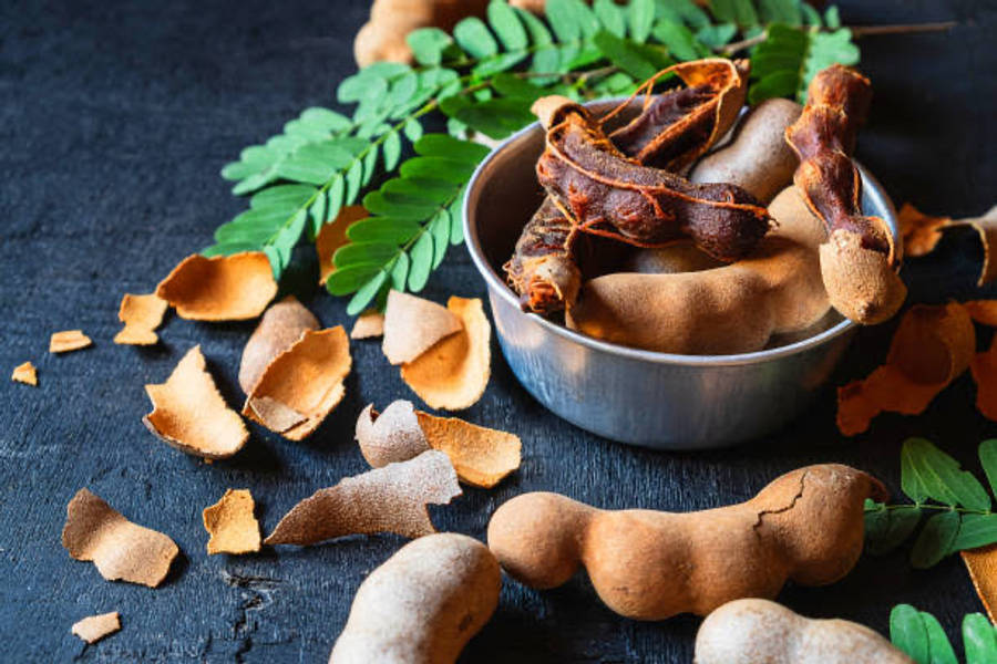 beans 桌布 壁紙 — 一個質樸的金屬碗盛裝著乾羅望子莢和散落的殼，放在深色紋理表面上，搭配新鮮綠葉。