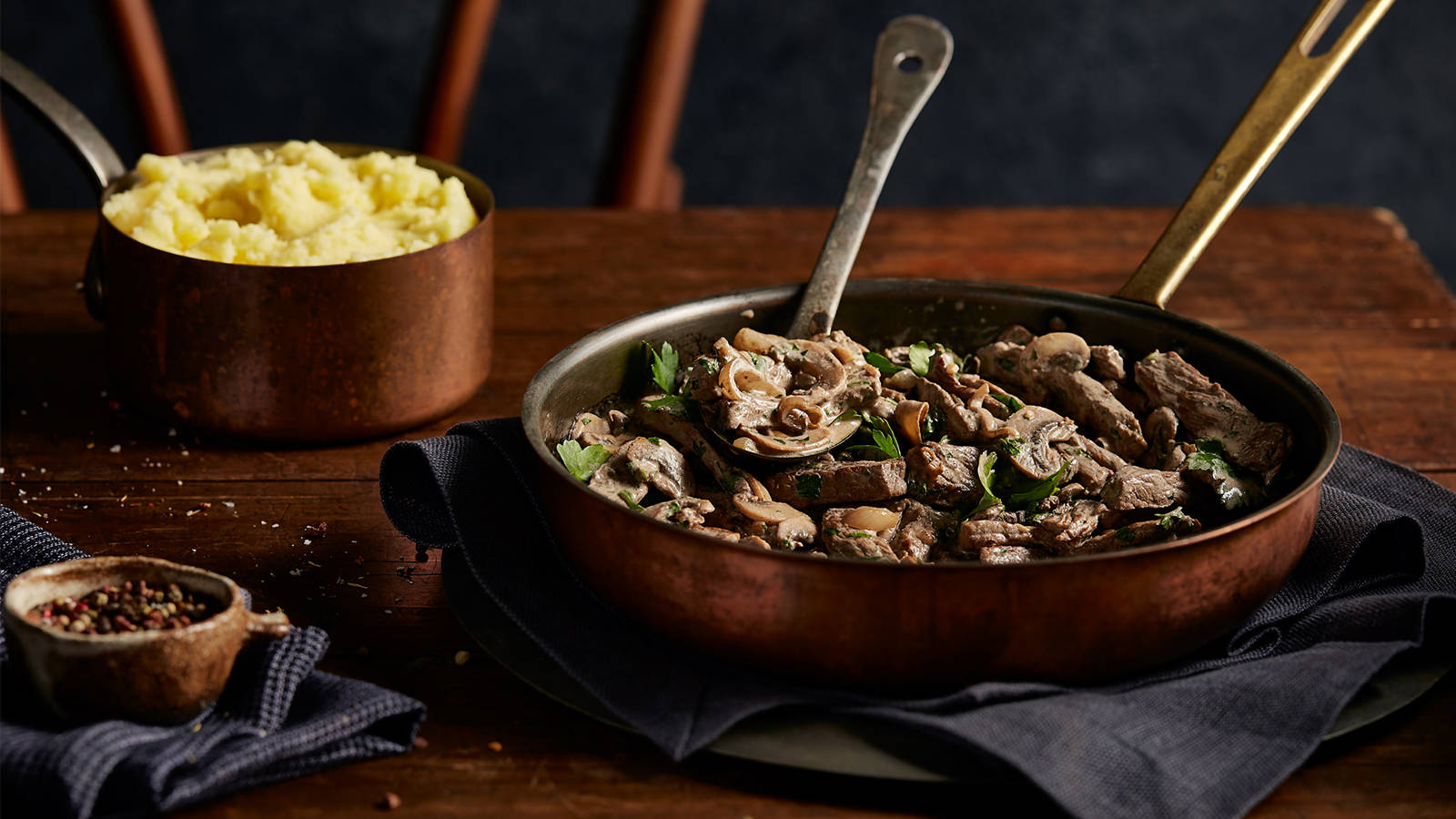 Beef Stroganoff 桌布 壁紙 — 一個質樸的銅鍋裝滿嫩牛肉和蘑菇，放在深色布上，旁邊有一鍋奶油馬鈴薯泥在木桌上。