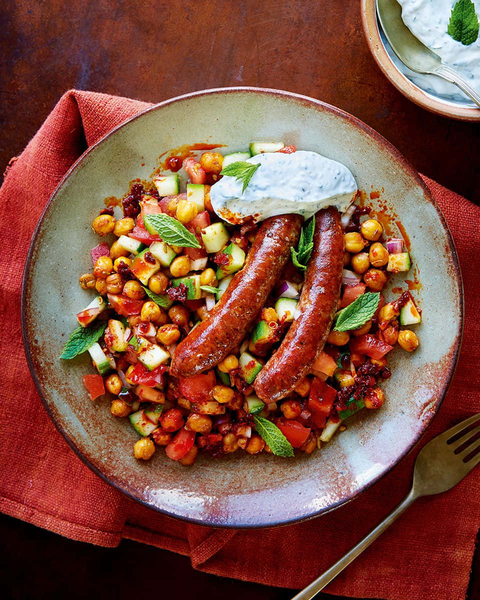 Sausage 桌布 壁紙 — 一個質樸的陶瓷碗盛裝著兩條香腸，放在充滿活力的鷹嘴豆沙拉中，搭配切丁蔬菜和薄荷，上面淋上優格醬，置於紅布上。