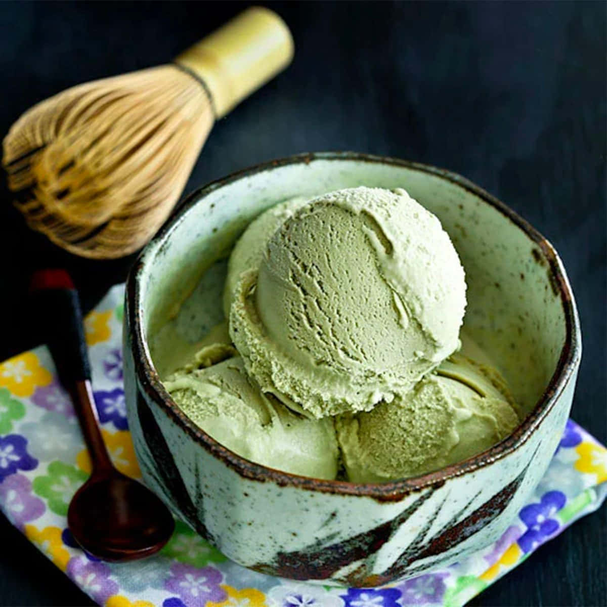 Japanese Sweets 桌布 壁紙 — 一個質樸的陶瓷碗盛載著 creamy 的綠茶冰淇淋，旁邊放著竹拂和木匙，置於花卉餐巾上。