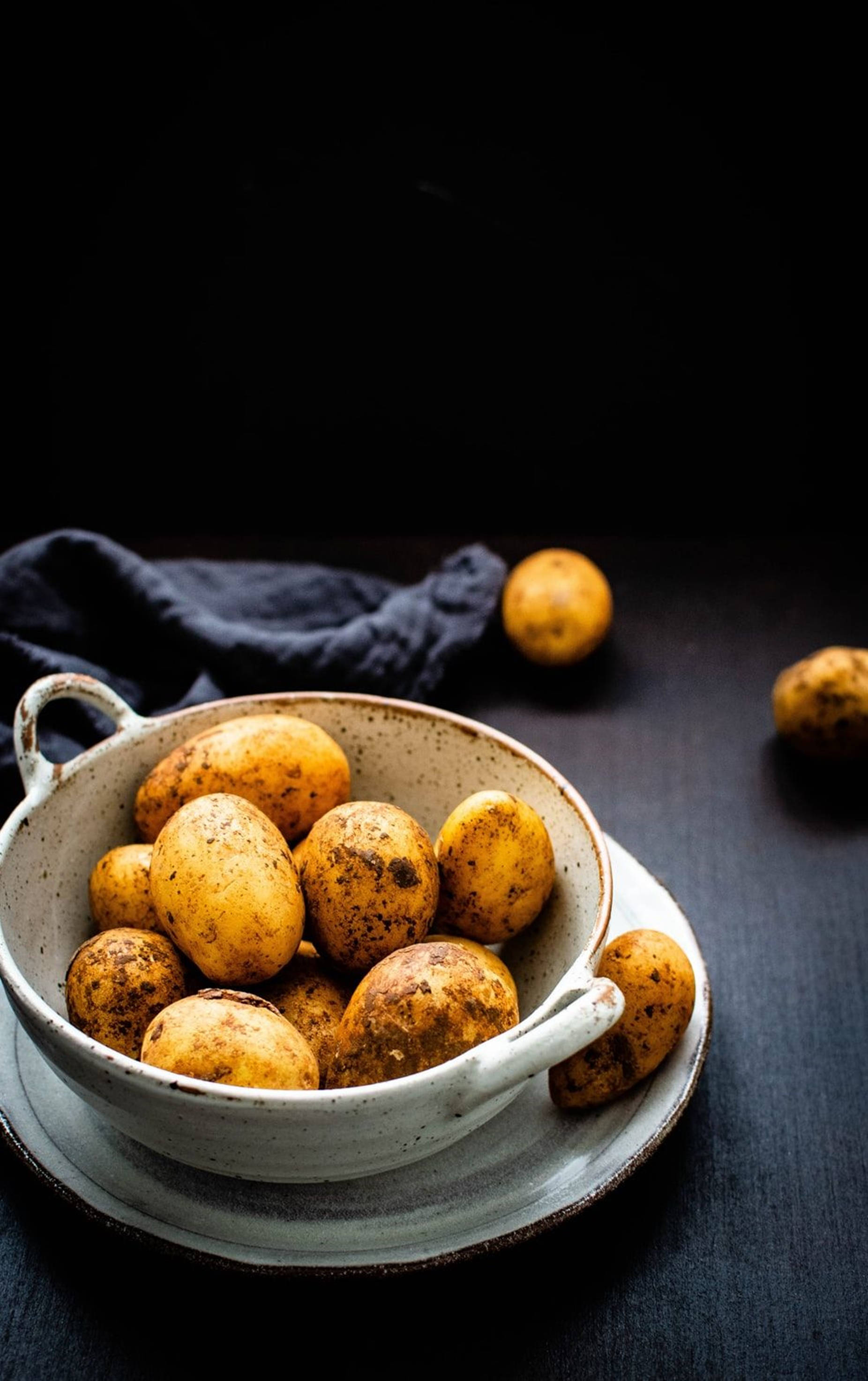 Potato 桌布 壁紙 — 一個質樸的陶瓷碗裝滿了土黃色的馬鈴薯，放在深色木質表面上，附近有一塊海軍藍色的布。