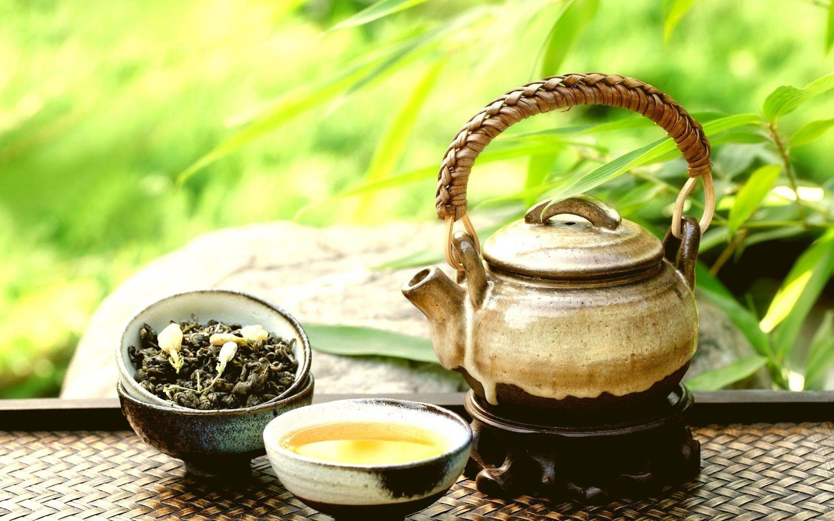 Garden 桌布 壁紙 — 一個質樸的陶瓷茶壺和茶杯放在編織墊上，旁邊有散裝茶葉，背景是柔焦的茂密綠竹。