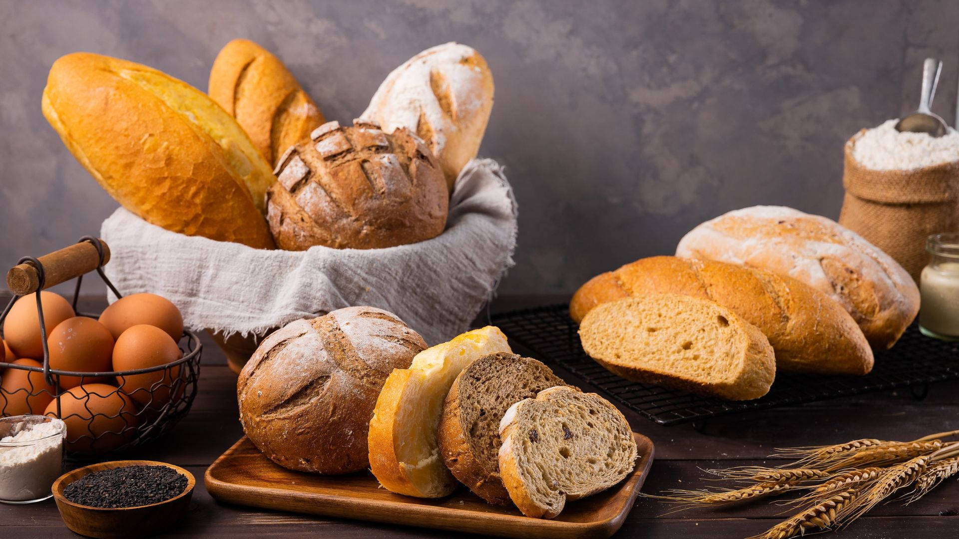 Bread 桌布 壁紙 — 一個質樸的靜物擺設，包含各種手工麵包、雞蛋和小麥，排列在木桌上，背景為有紋理的灰色。