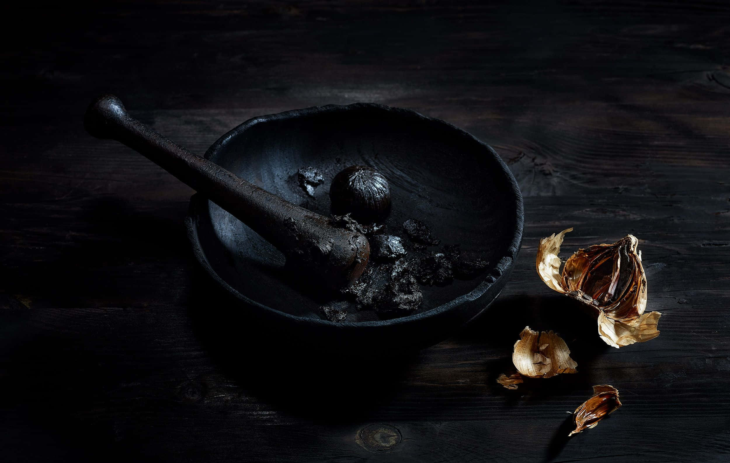 Black Garlic 桌布 壁紙 — 一個質樸的黑色研缽和研杵，裡面有壓碎的大蒜，放在深色木桌上，旁邊有乾蒜瓣，在戲劇性的燈光下。