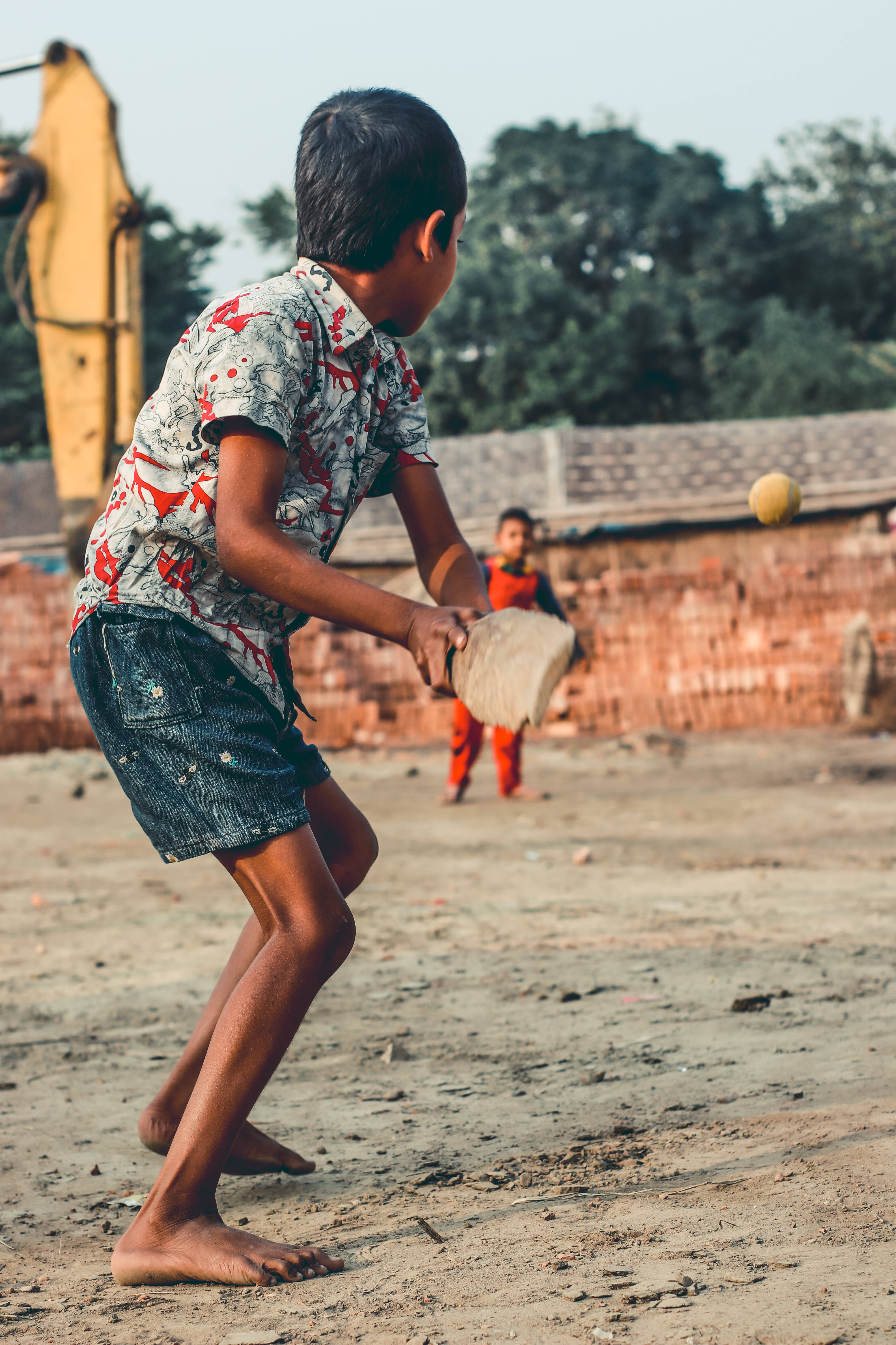 Bangladesh 桌布 壁紙 — 一個赤腳男孩穿著圖案襯衫，在塵土飛揚的院子裡揮動球拍擊球，身後有磚牆和樹木。