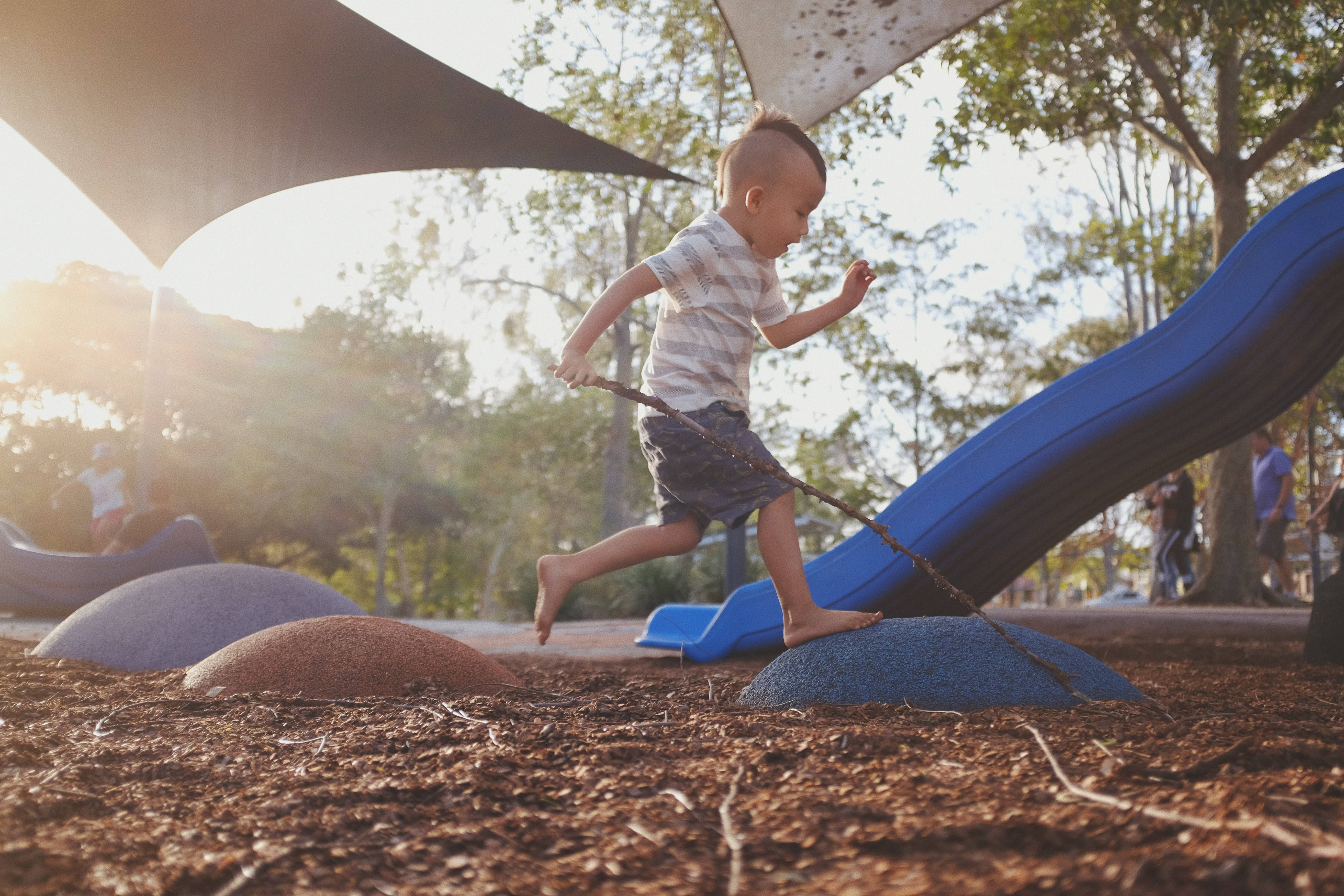 Playground 桌布 壁紙 — 一個赤腳的幼兒在明亮的陽光下，手裡拿著樹枝，充滿活力地跑過遊樂場。