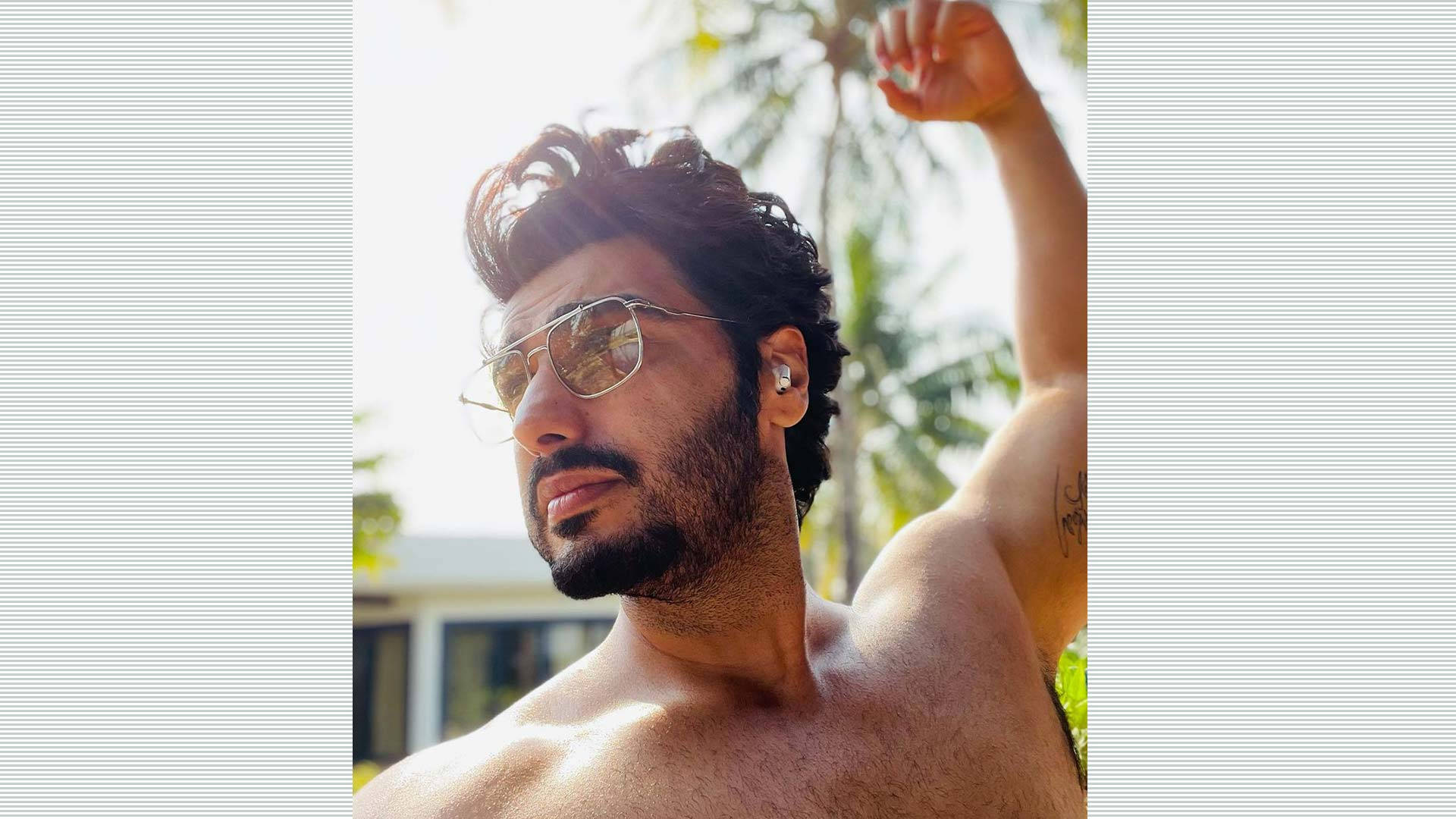 Arjun Kapoor 桌布 壁紙 — 一個赤膊、深色頭髮和鬍鬚的男人，戴著飛行員太陽眼鏡，在棕櫚樹的明亮熱帶背景下舉起手臂。