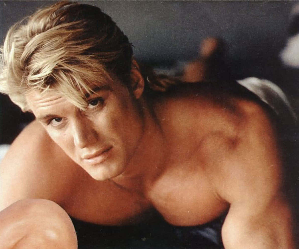 Dolph Lundgren 桌布 壁紙 — 一個赤膊、肌肉發達、金髮蓬鬆的男人在昏暗的房間裡向前傾，專注地凝視著鏡頭。