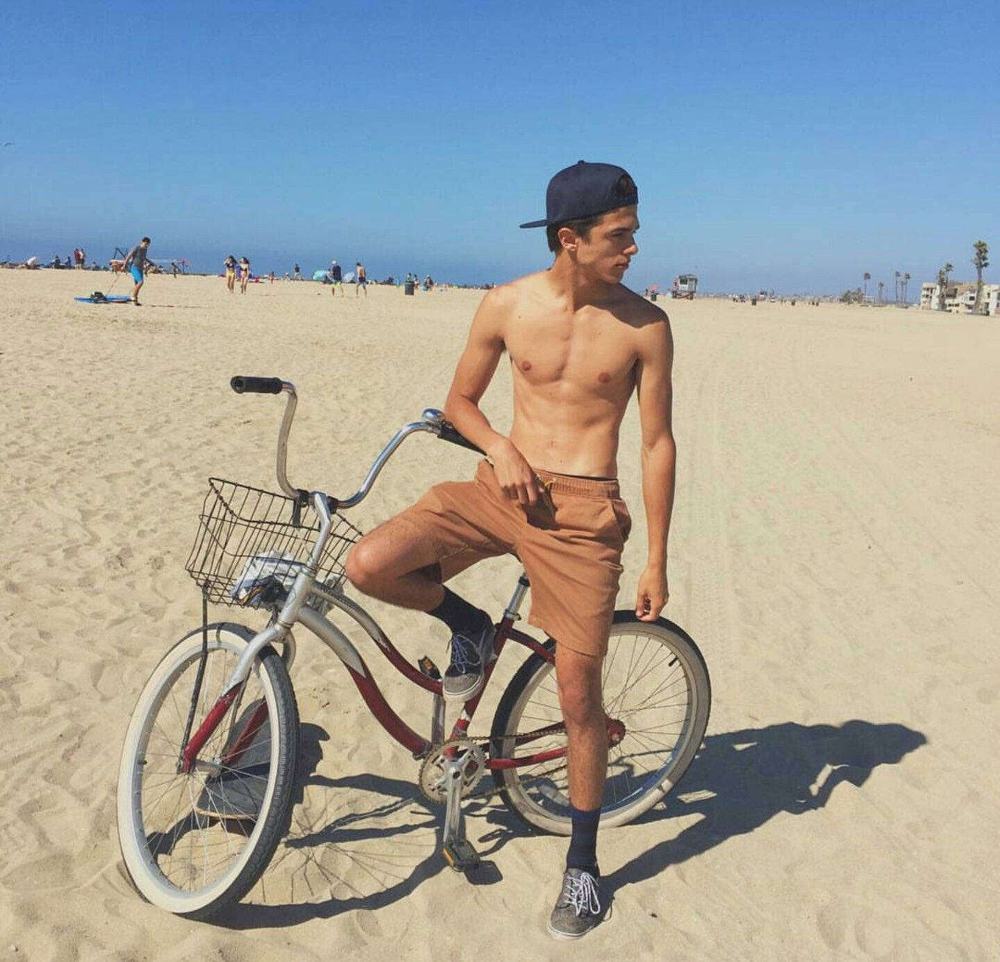 Rivera 桌布 壁紙 — 一個赤膊戴帽的年輕男子，在陽光明媚的沙灘上隨意靠在一輛復古沙灘巡洋艦腳踏車上。