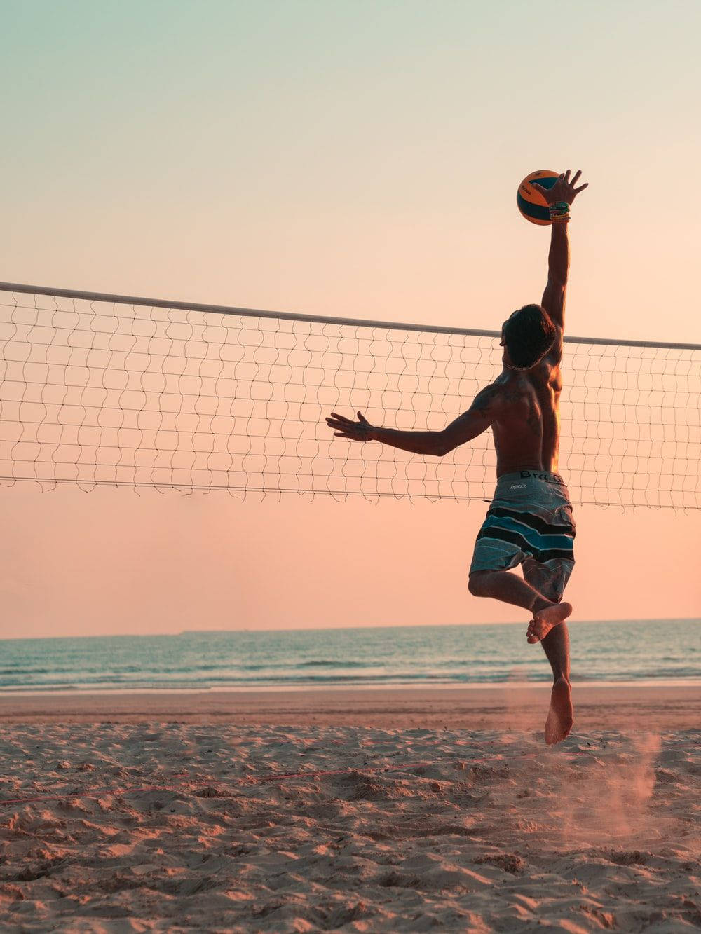 Volleyball Aesthetic 桌布 壁紙 — 一個赤膊男子在日落時分的沙灘上高高跳起，將排球扣過網，身後是海浪。