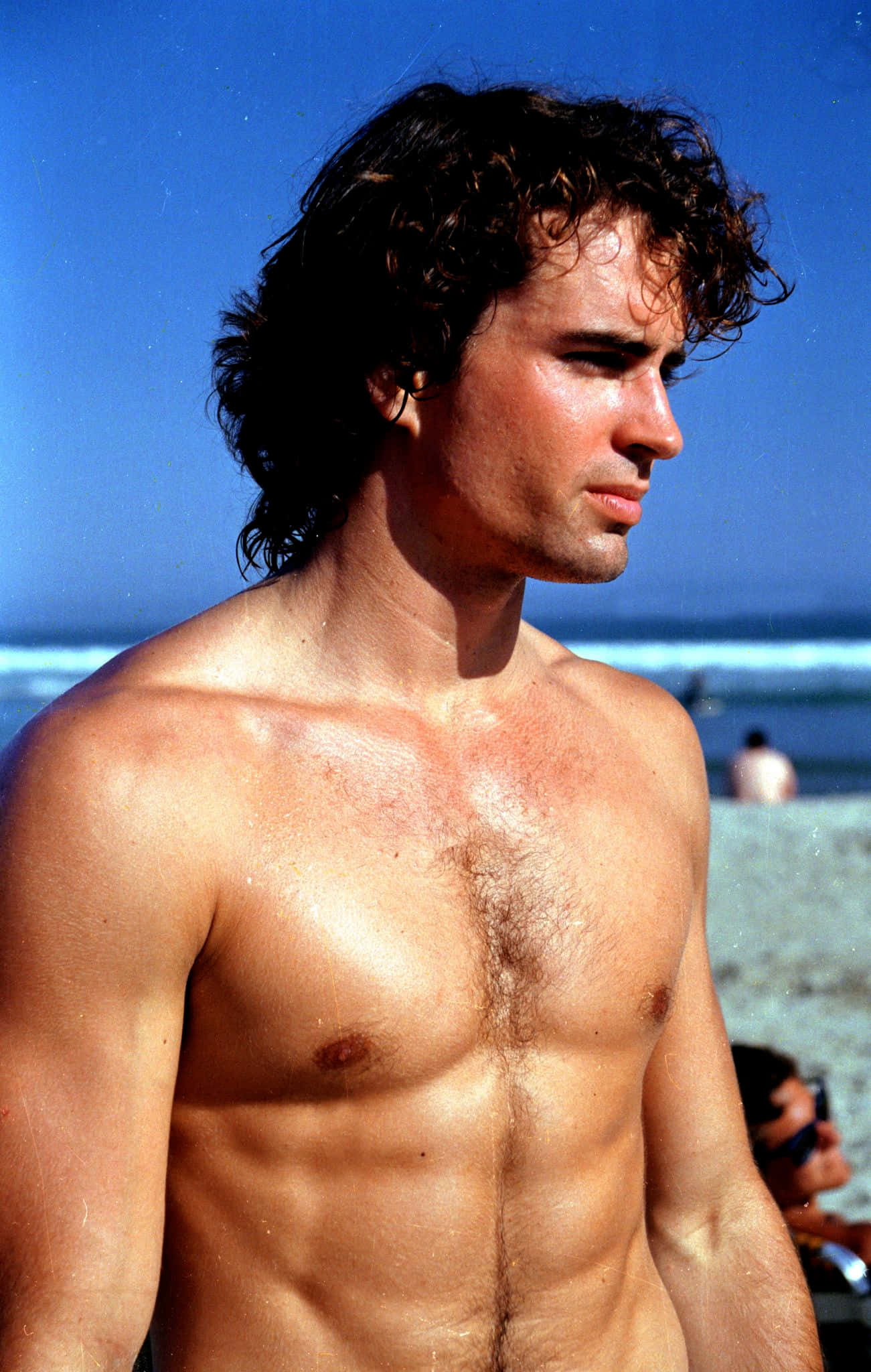 Jason Patric 桌布 壁紙 — 一個赤膊的男人，濕漉漉的捲髮，站在陽光普照的沙灘上，在晴朗的藍天下凝視著海洋。