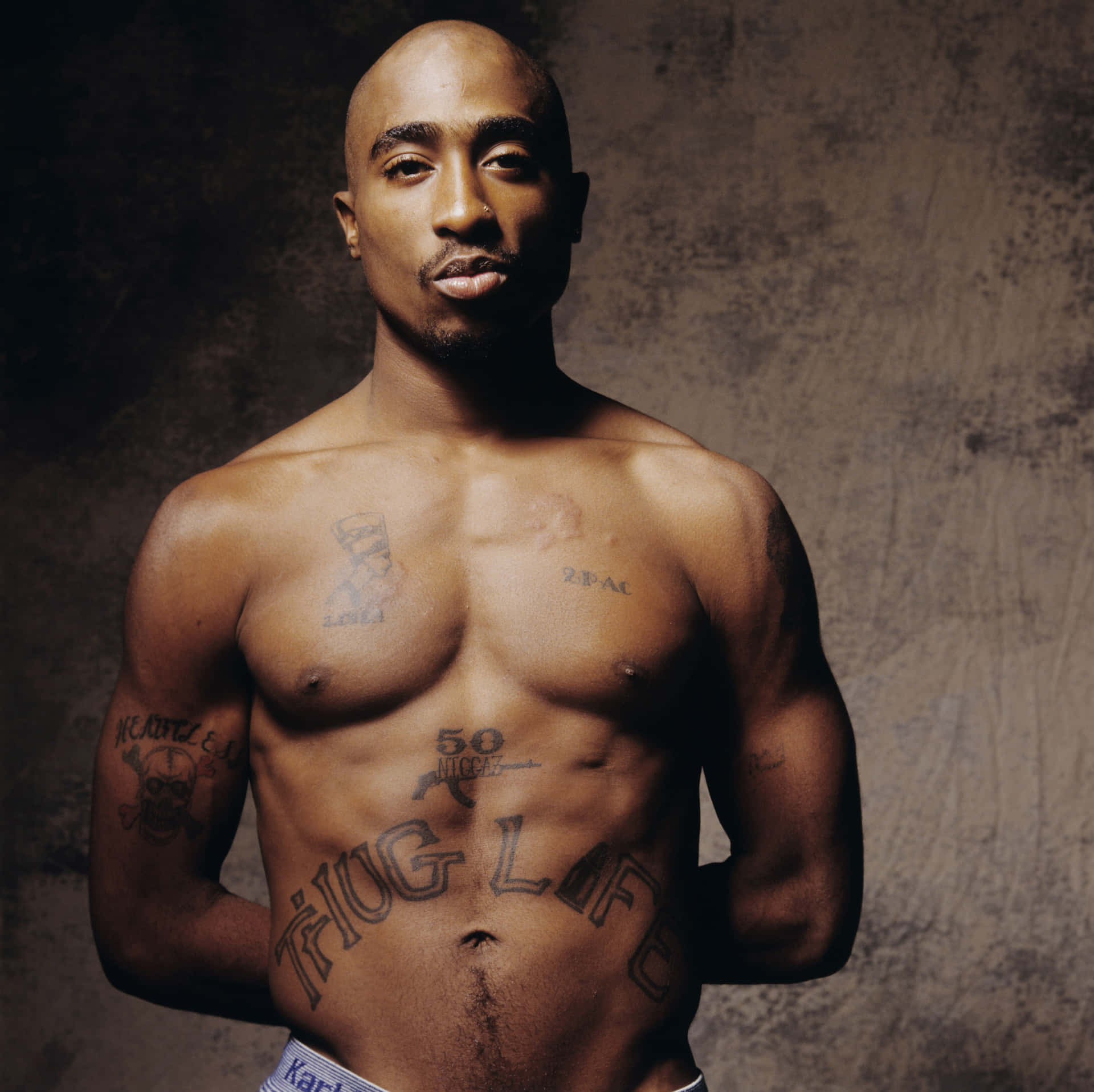 tupac 桌布 壁紙 — 一個赤膊的男人，身上有刺青，包括腹部的「THUG LIFE」，站在有紋理的棕色牆前面。