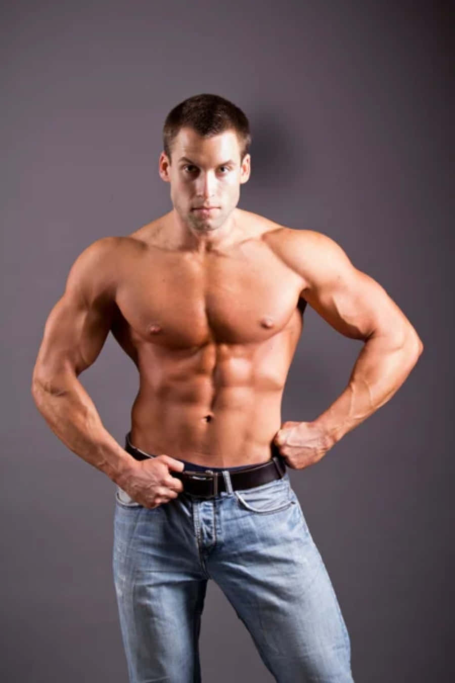 Muscle 桌布 壁紙 — 一個赤膊的肌肉男，擁有明顯的腹肌和手臂，自信地穿著牛仔褲站在純灰色工作室背景前。