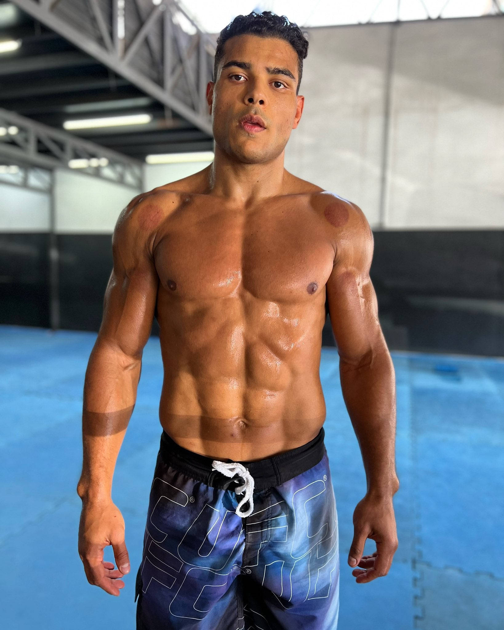 Fitness 桌布 壁紙 — 一個赤膊肌肉男，腹肌線條分明，自信地站在健身房的藍色墊子上，周圍是工業風照明。