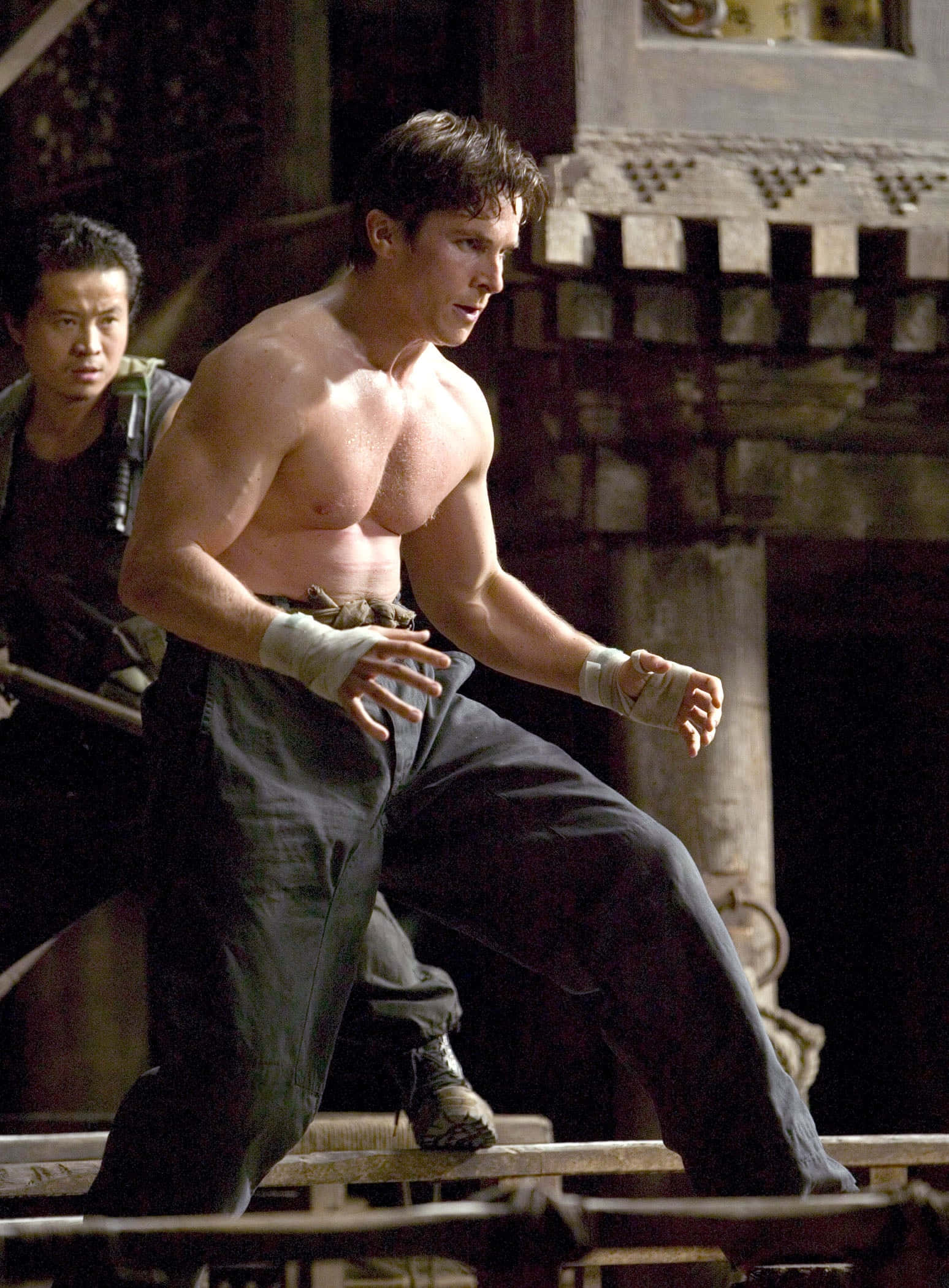 Batman Begins 桌布 壁紙 — 一個赤膊肌肉男以纏手戰鬥姿勢站在昏暗古廟內的木樑上。