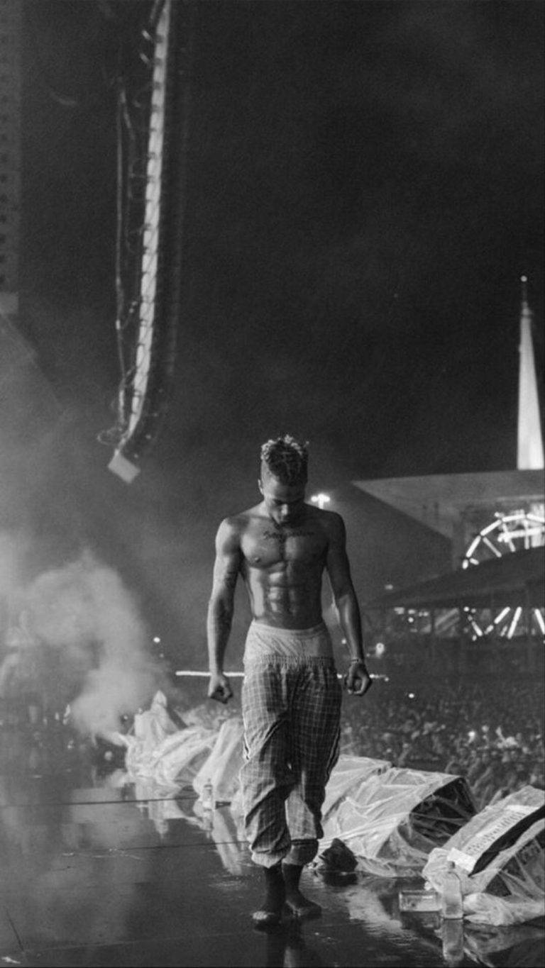 xxxtentacion 美學 桌布 壁紙 — 一個赤膊肌肉男赤腳走過煙霧，在夜燈下的濕滑舞台上，下方有群眾。