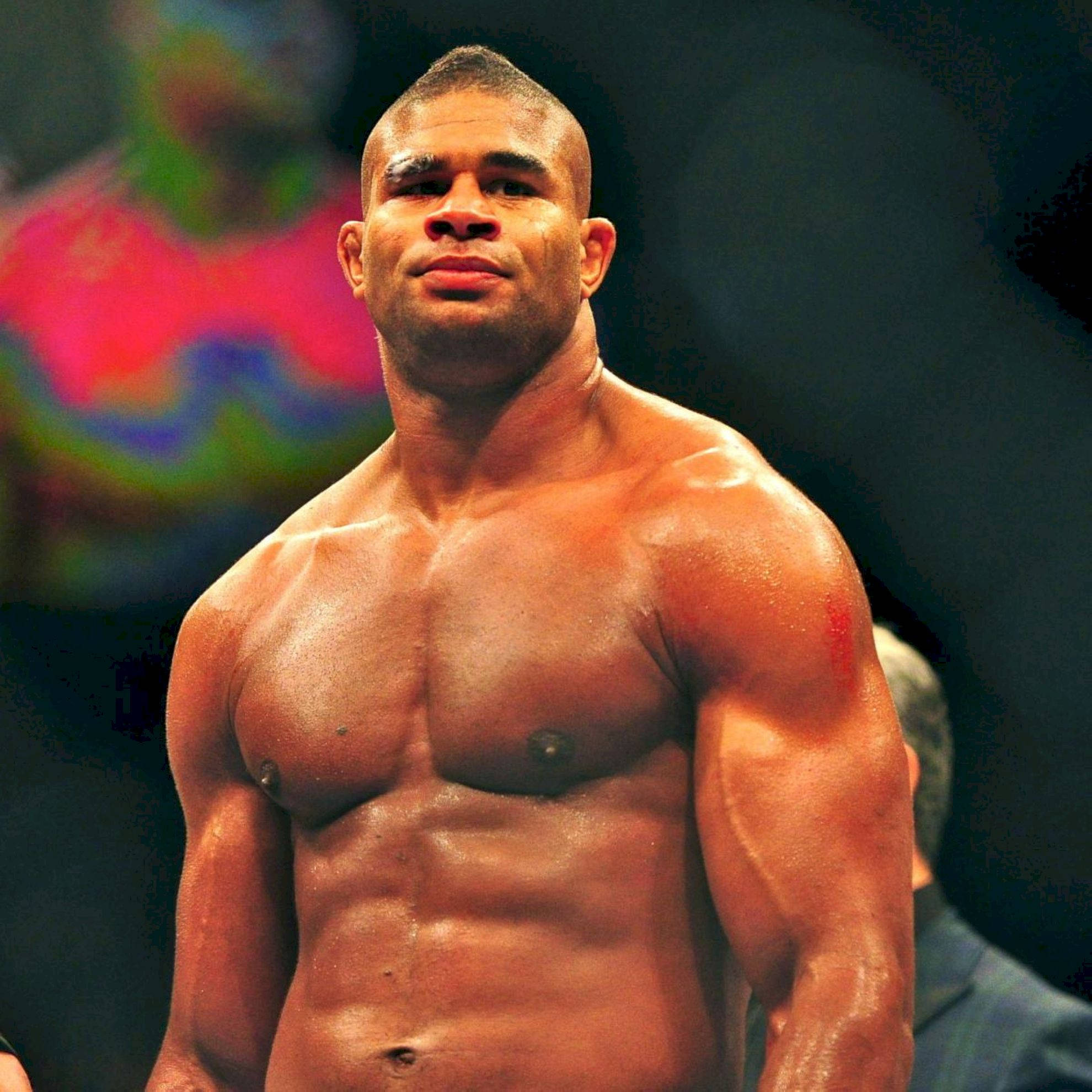 UFC 桌布 壁紙 — 一個赤膊肌肉發達的戰士，神情專注，站在明亮的競技場燈光下，背景是模糊的彩色。