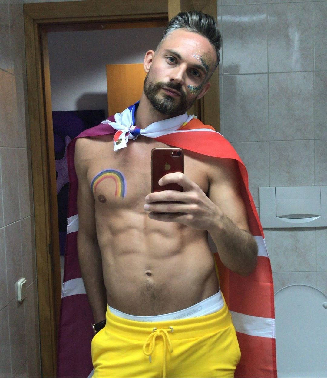 Bisexual 桌布 壁紙 — 一個赤裸上身、帶有彩虹刺青和旗幟斗篷的男人在磁磚浴室裡對著鏡子自拍。