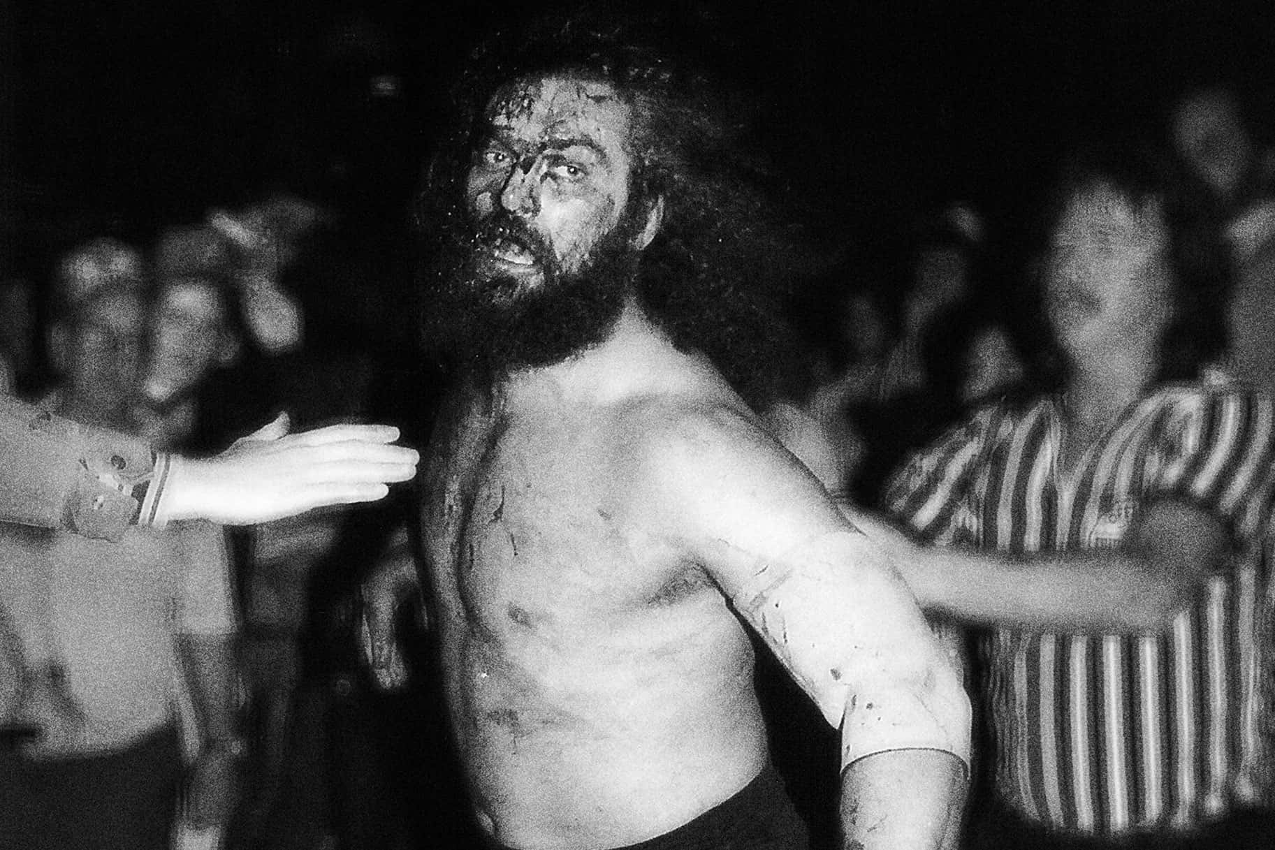 WWE 桌布 壁紙 — 一個赤裸上身、鬍鬚滿面、渾身泥濘的男人站在舞台中央，人們在粗糙的黑白照片中擠在他周圍。