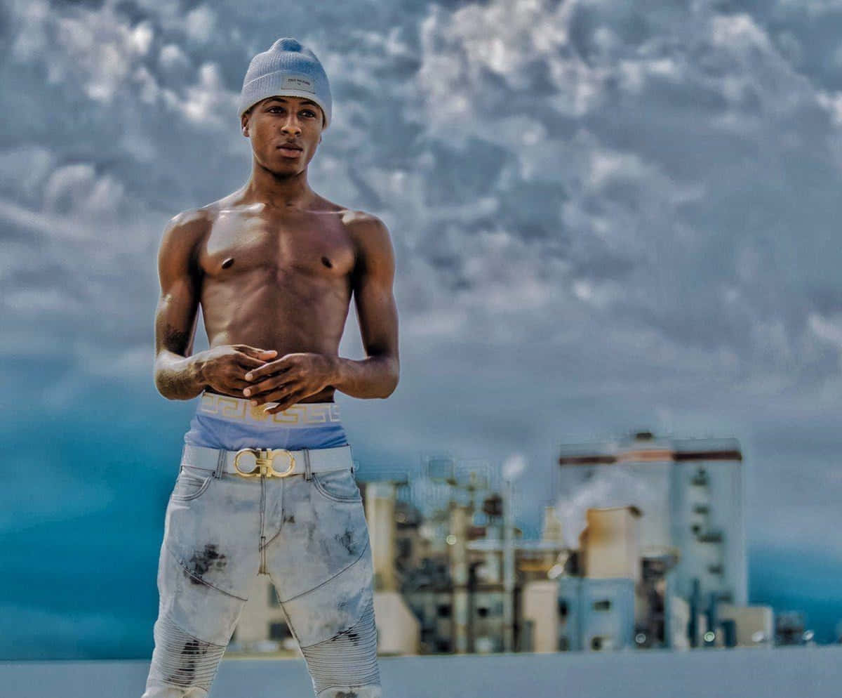 NBA YoungBoy 桌布 壁紙 — 一個赤裸上身的年輕男子戴著毛帽和破損牛仔褲，自信地站在戲劇性的多雲天空下，背景是工業建築。