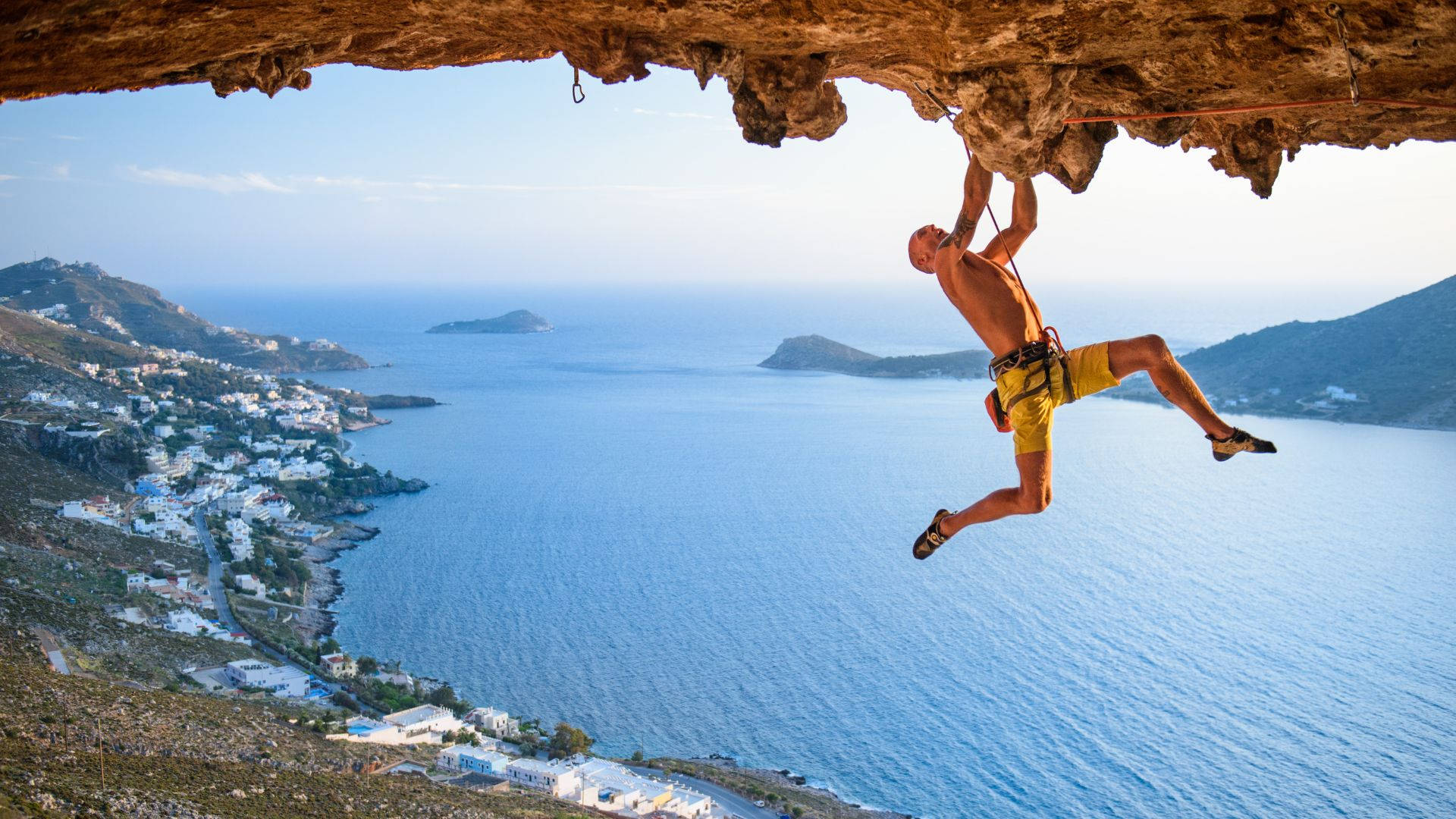Climbing 桌布 壁紙 — 一個赤裸上身的攀岩者懸掛在懸崖上，下方是碧綠色的海洋和白色村莊，天空明亮。