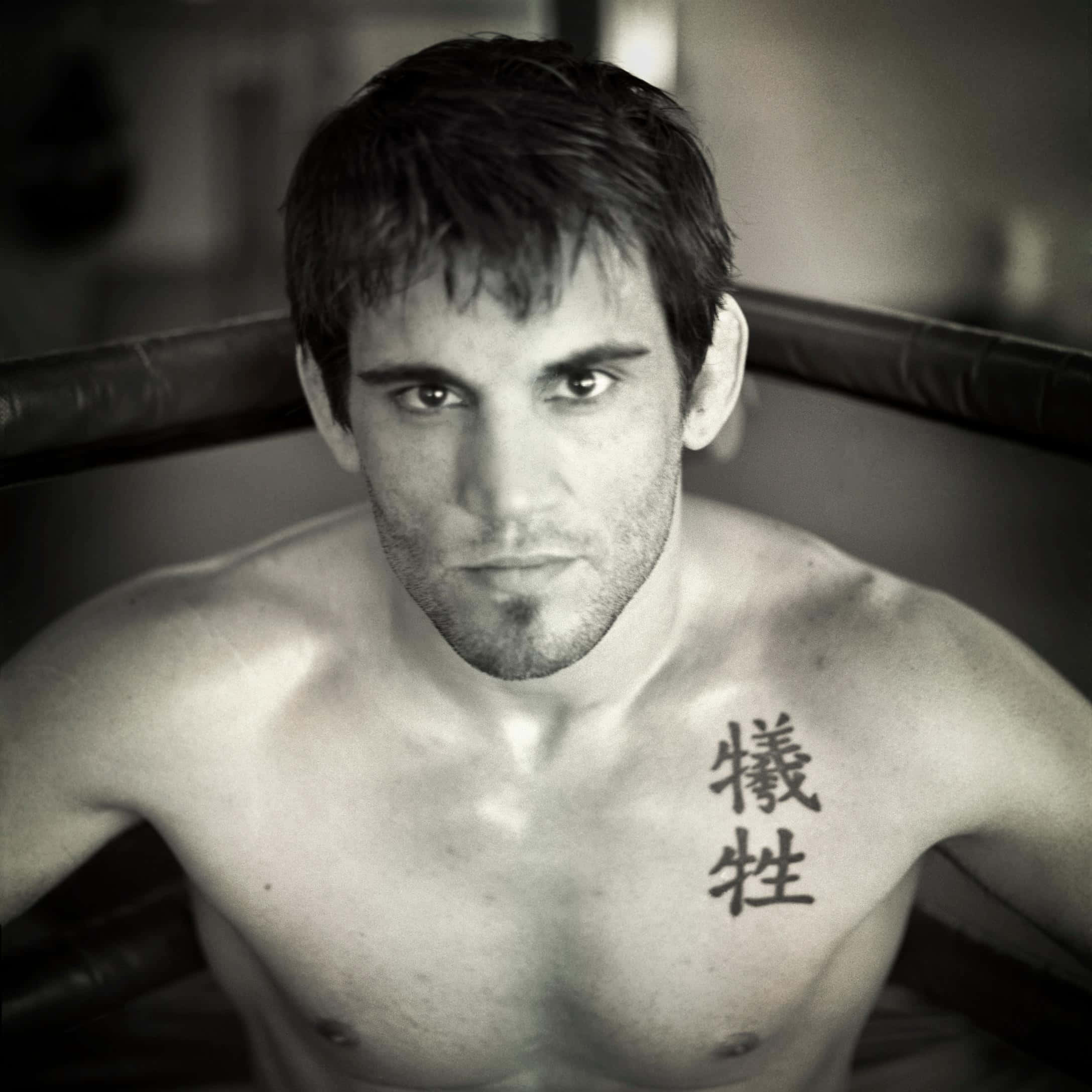 UFC 桌布 壁紙 — 一個赤裸上身的男人，帶著嚴厲的眼神，胸前有一個中文刺青，站在黑白拳擊場內。