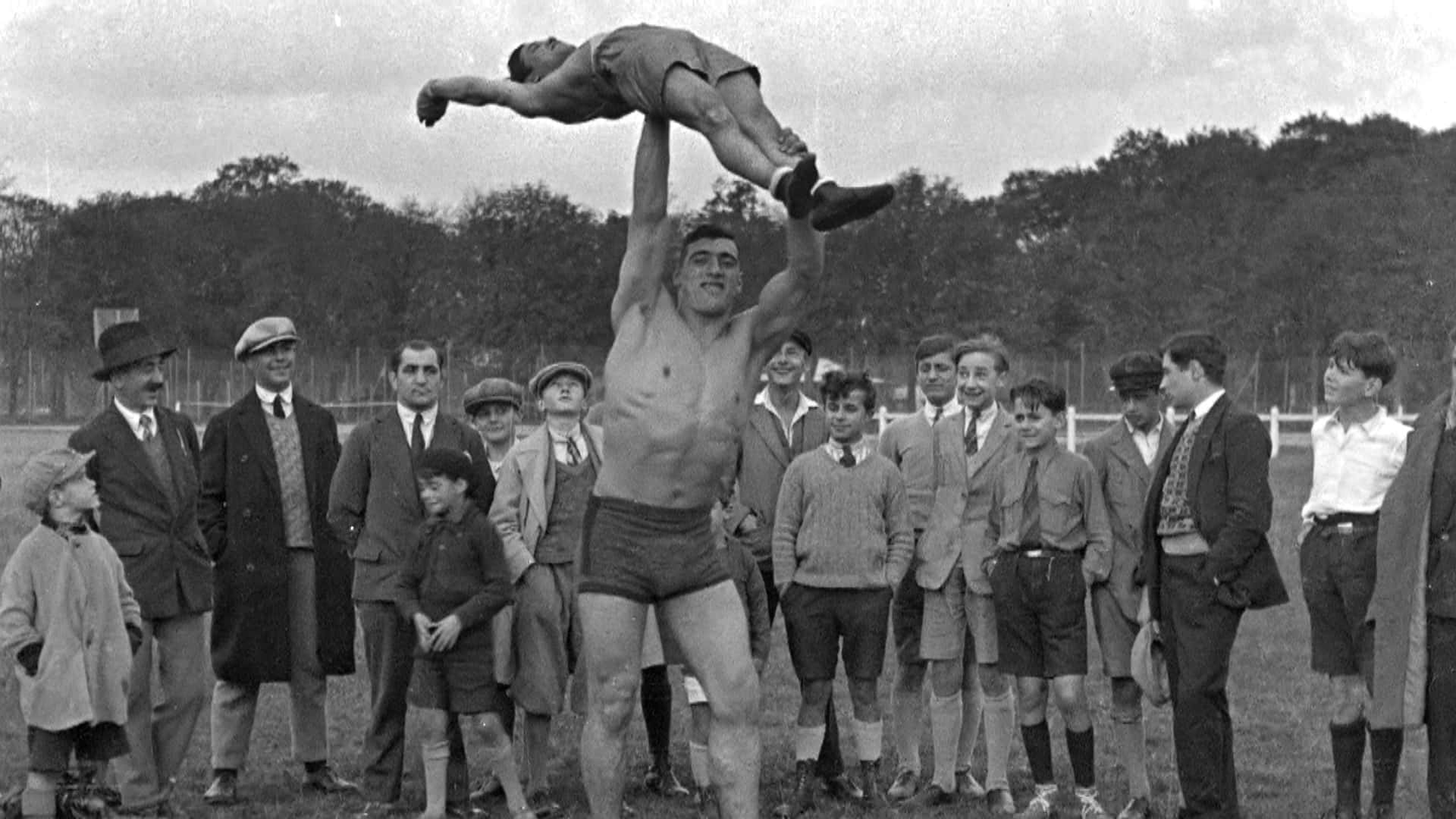 boxer 桌布 壁紙 — 一個赤裸上身的男人在草地上將另一個男人舉過頭頂，一群人在旁觀看，復古黑白照片。