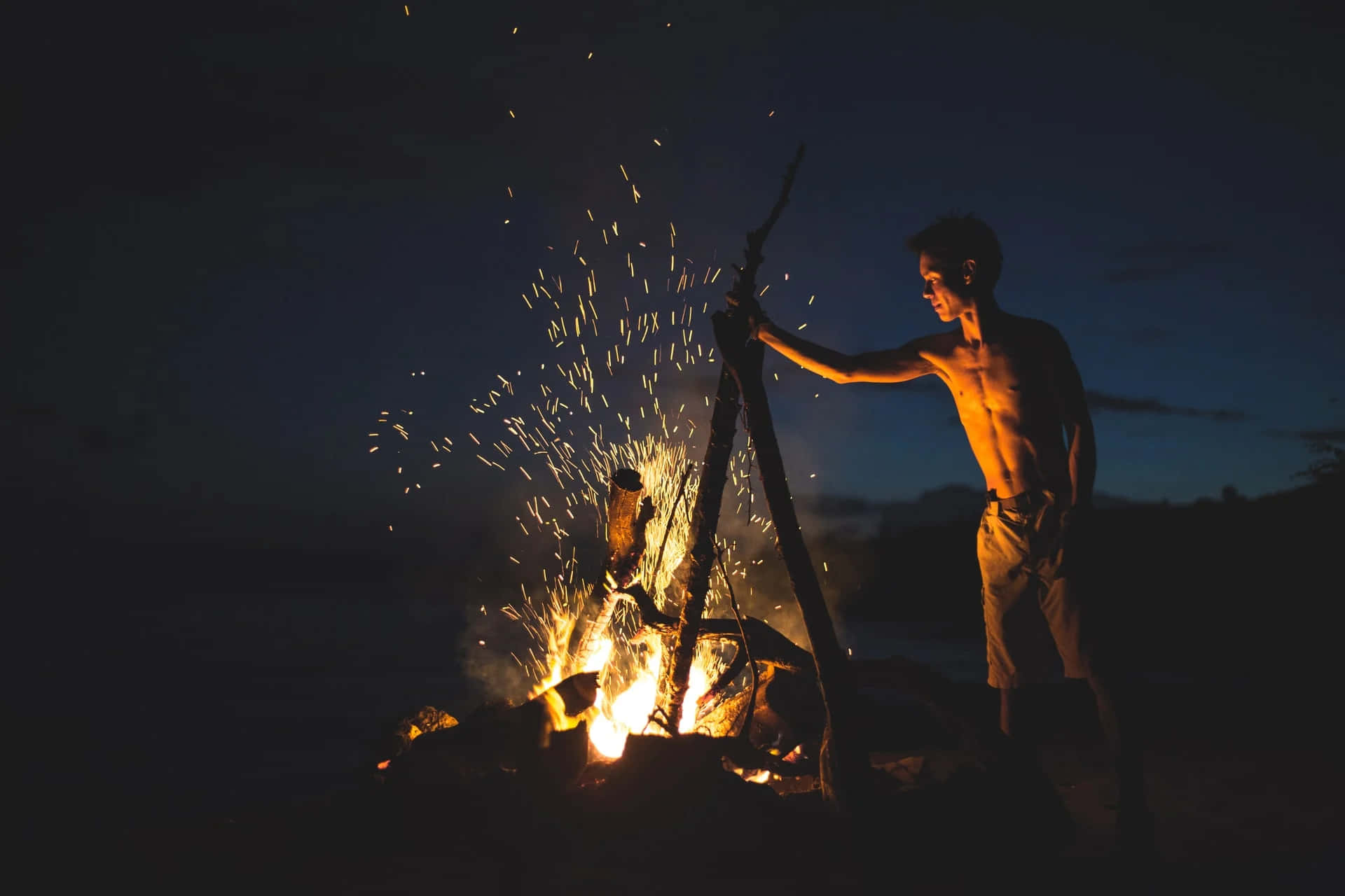 Bonfire 桌布 壁紙 — 一個赤裸上身的男人在黃昏時攪動熊熊營火，金色火花在深藍色暮色天空中飛舞。