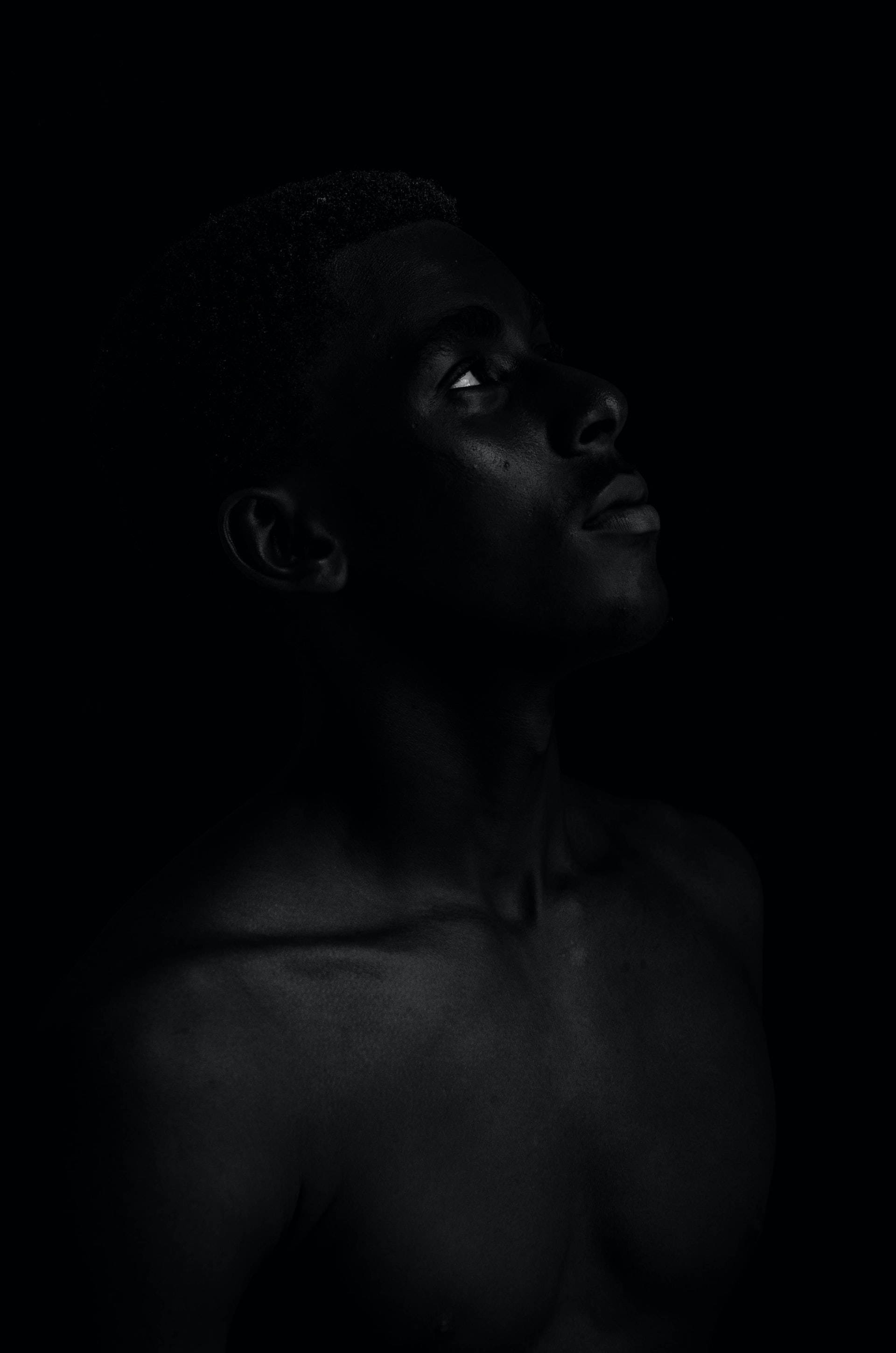 black aesthetic tumblr iPhone 桌布 壁紙 — 一個赤裸上身的男人的臉和肩膀，在純黑色背景上從上方戲劇性地照亮，突顯他向上凝視的目光和清晰的面部特徵。