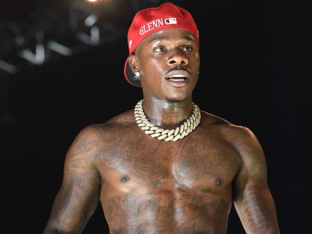 Dababy 桌布 壁紙 — 一個赤裸上身的男子，身上有刺青，戴著紅色帽子和粗金項鍊，背景為深色。
