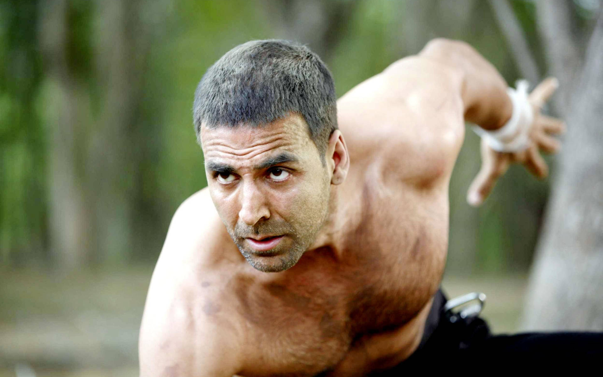 Akshay Kumar 桌布 壁紙 — 一個赤裸上身的肌肉男，在模糊的森林背景下擺出動態武術姿勢，神情專注。