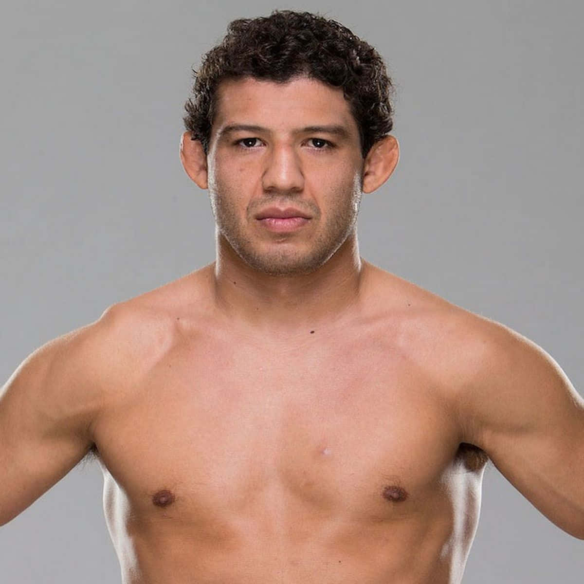 UFC 桌布 壁紙 — 一個赤裸上身的肌肉男，留著捲曲的深色頭髮，在純灰色的攝影棚背景前，專注地凝視著鏡頭。