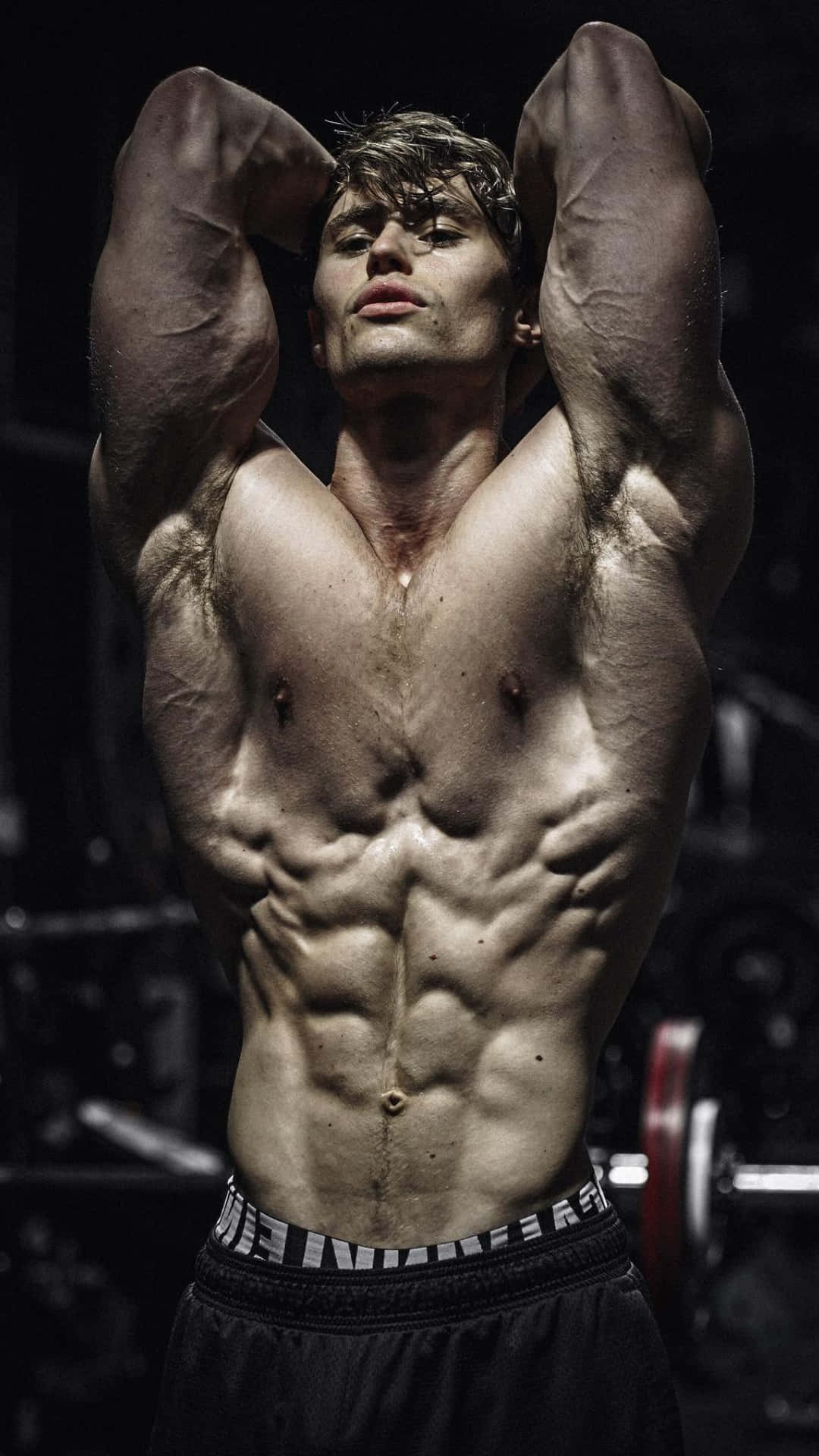 Fitness 桌布 壁紙 — 一個赤裸上身的肌肉男在光線昏暗的健身房中繃緊手臂和軀幹，背景有啞鈴。