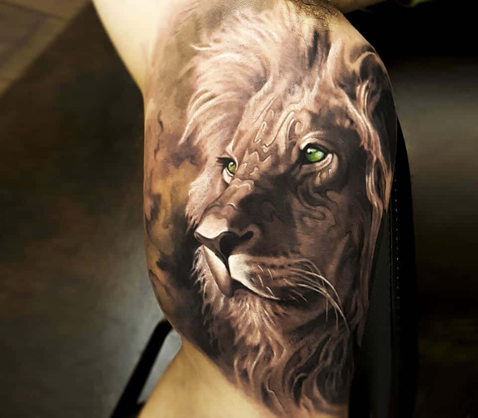 Lion Head 桌布 壁紙 — 一個超寫實的獅子刺青，帶有發光的綠色眼睛，以細緻的陰影覆蓋了一個人的肩膀和上臂。