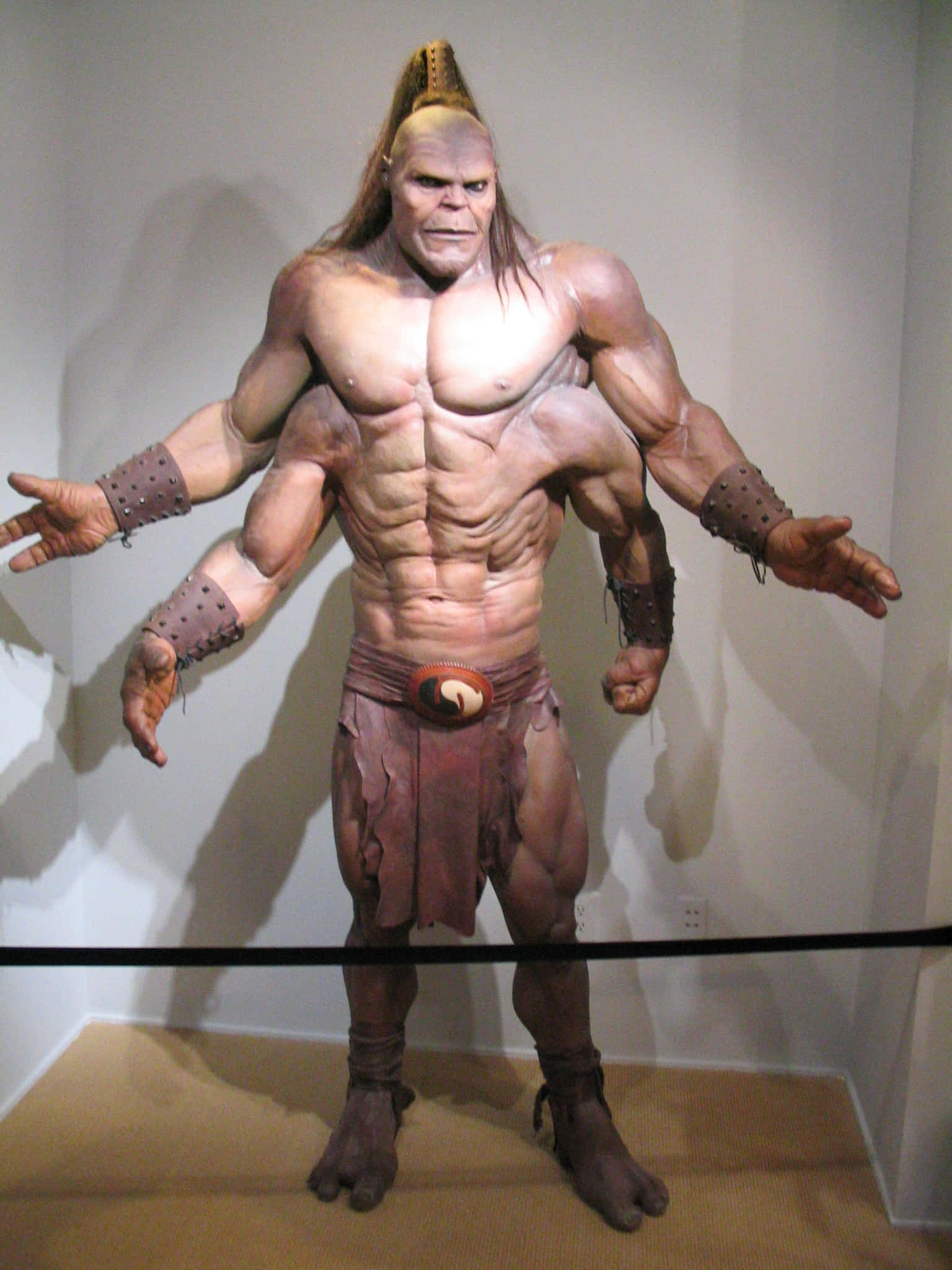 Mortal Kombat Goro 桌布 壁紙 — 一個超寫實的蠟像，描繪一位肌肉發達、紮馬尾辮、身穿部落裝備的戰士，站立在博物館的展示櫃中。