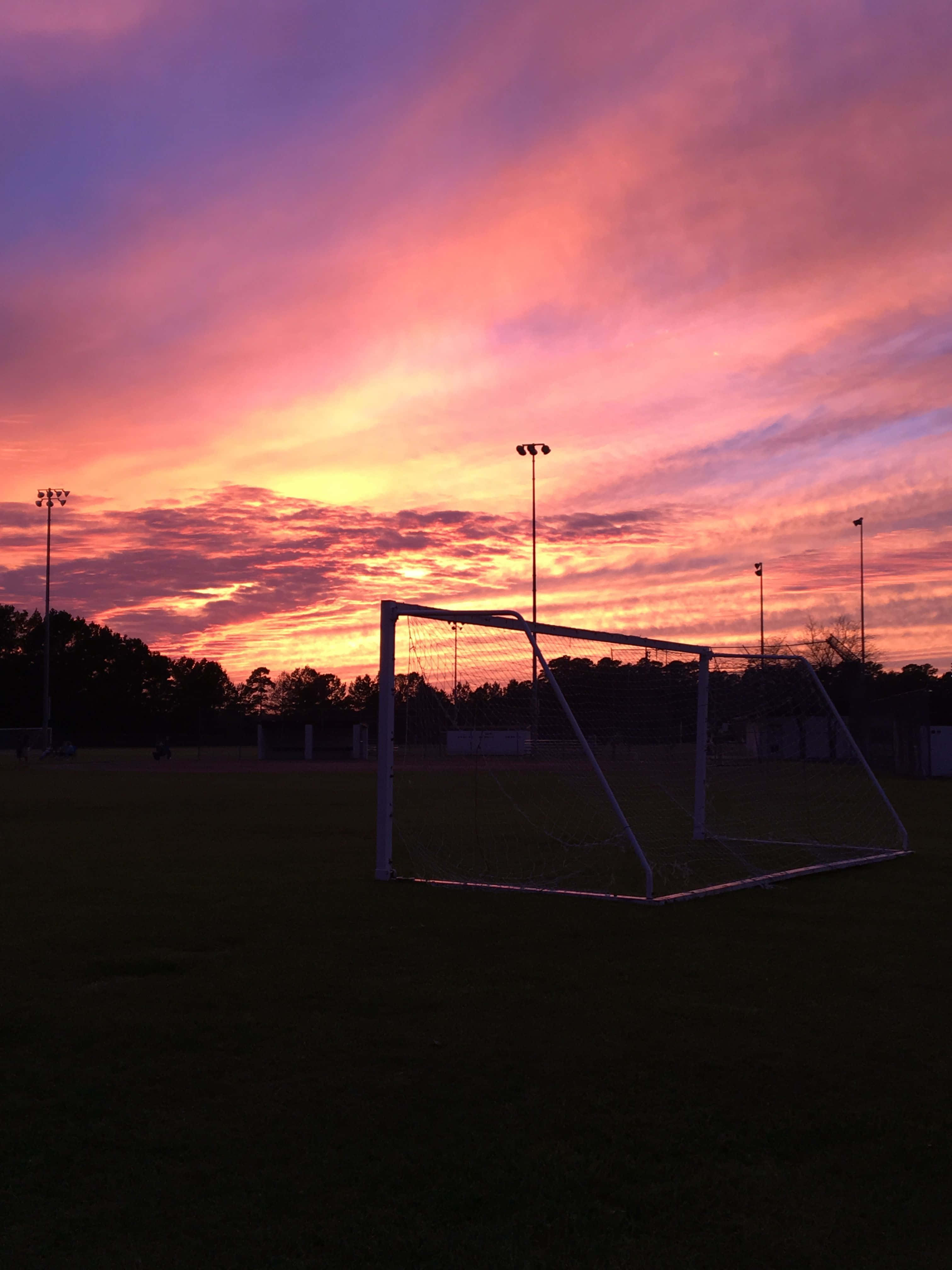 Soccer Aesthetic 桌布 壁紙 — 一個足球龍門在夕陽映照下形成剪影，天空被火熱的橙色、粉紅色和紫色雲彩染得色彩繽紛。