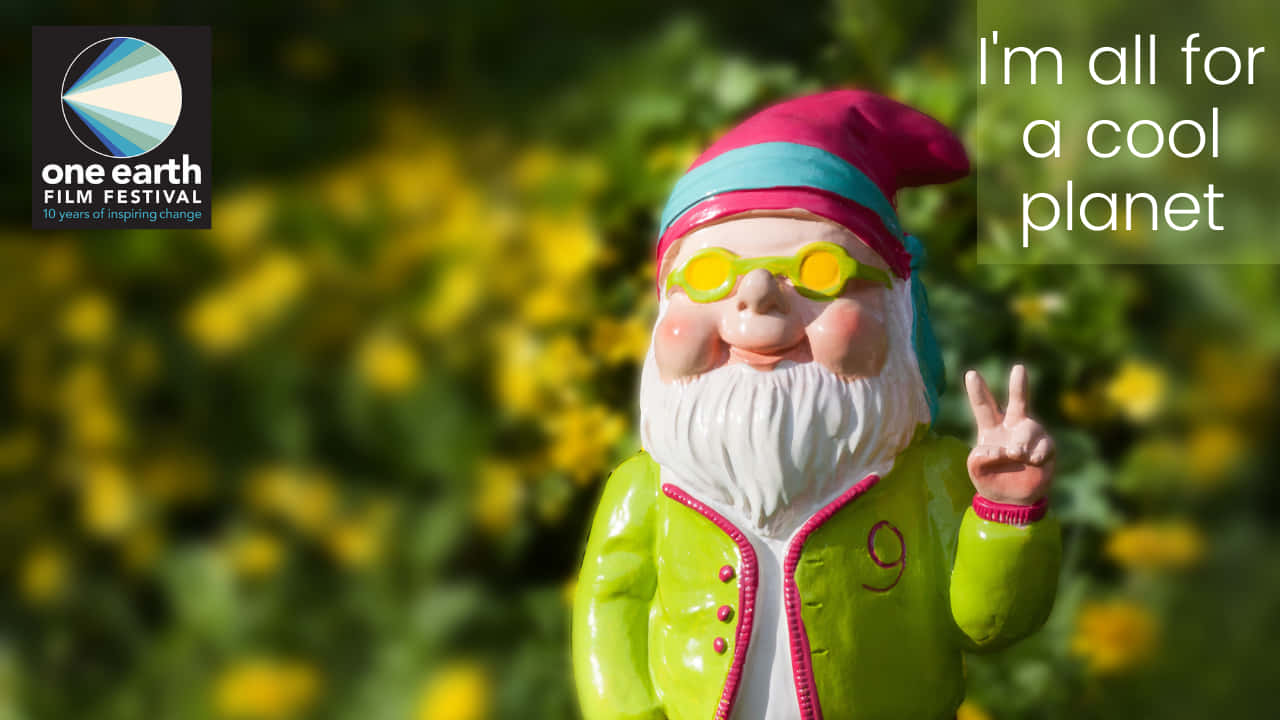 節日 桌布 壁紙 — 一個身穿亮綠色外套和粉紅色帽子的快樂花園小矮人，在模糊的黃色花朵背景前比出和平手勢。
