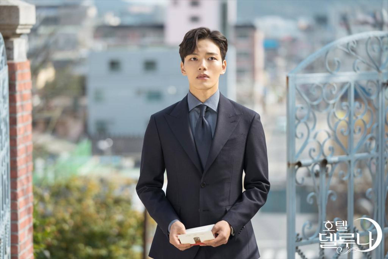 Jin 桌布 壁紙 — 一個身穿俐落深色西裝的男人站在陽台上，拿著一個盒子，在模糊的城市背景下若有所思地凝視著。