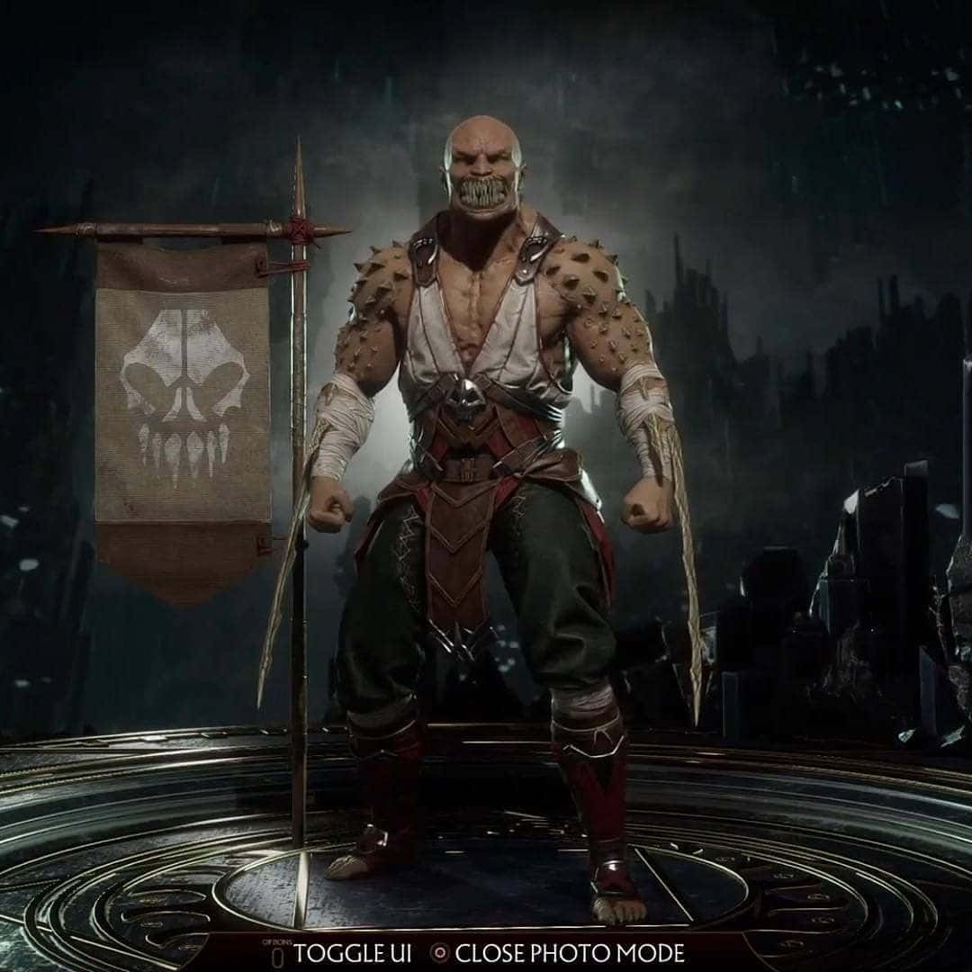 Mortal Kombat Baraka 桌布 壁紙 — 一個身穿尖刺盔甲、手持骷髏旗幟的威猛光頭戰士，在戲劇性燈光下，站在黑暗潮濕的廢墟中。