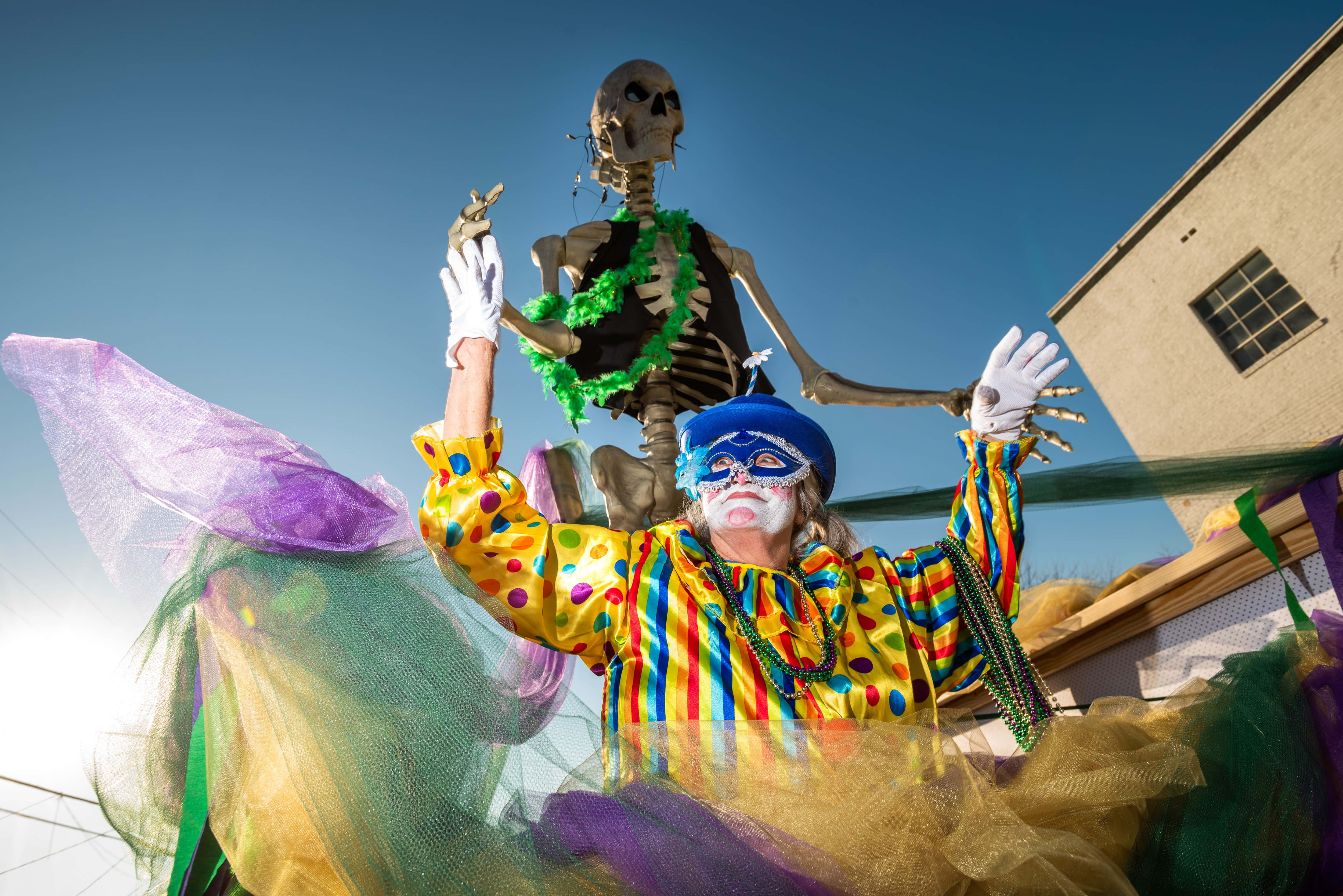 Mardi Gras 桌布 壁紙 — 一個身穿彩虹服飾、戴著面具的鮮豔小丑，站在一個飾有綠色珠子的骷髏人物下方，背景是清澈的藍天。