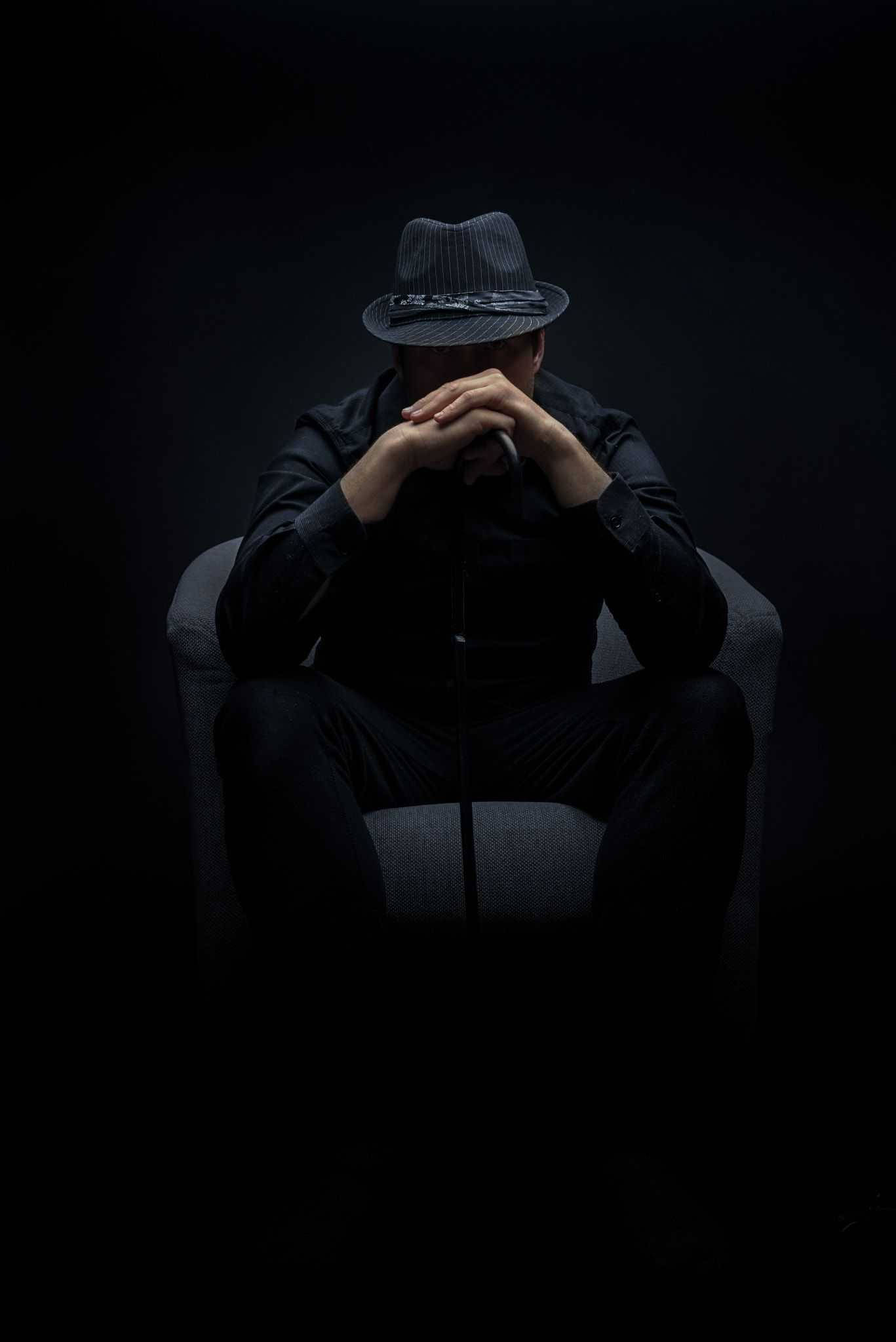 Mafia 桌布 壁紙 — 一個身穿斗篷、頭戴軟呢帽的身影蜷縮在陰影中，雙手交疊在臉上，坐在燈光昏暗的椅子上。