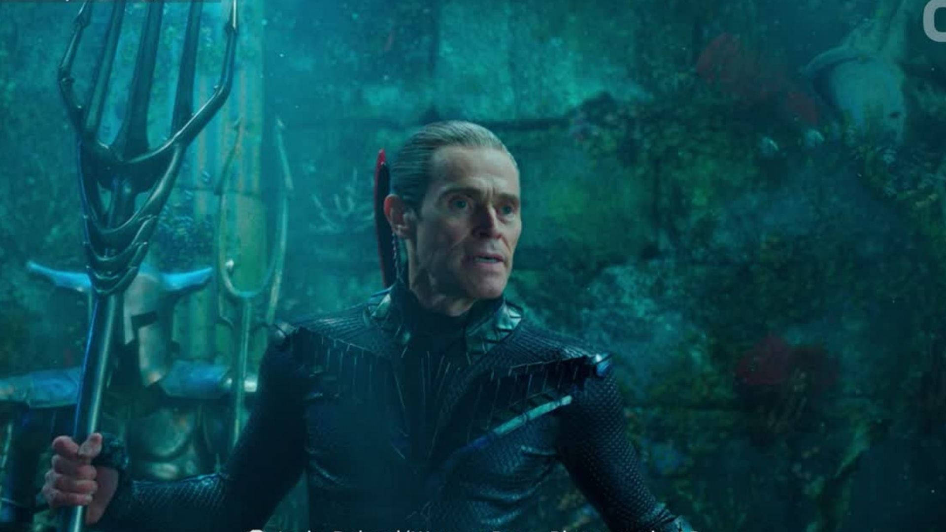 Willem Dafoe 桌布 壁紙 — 一個身穿暗色盔甲的男人在珊瑚和廢墟之間的水下拿著三叉戟，呈現青綠色的海洋場景。