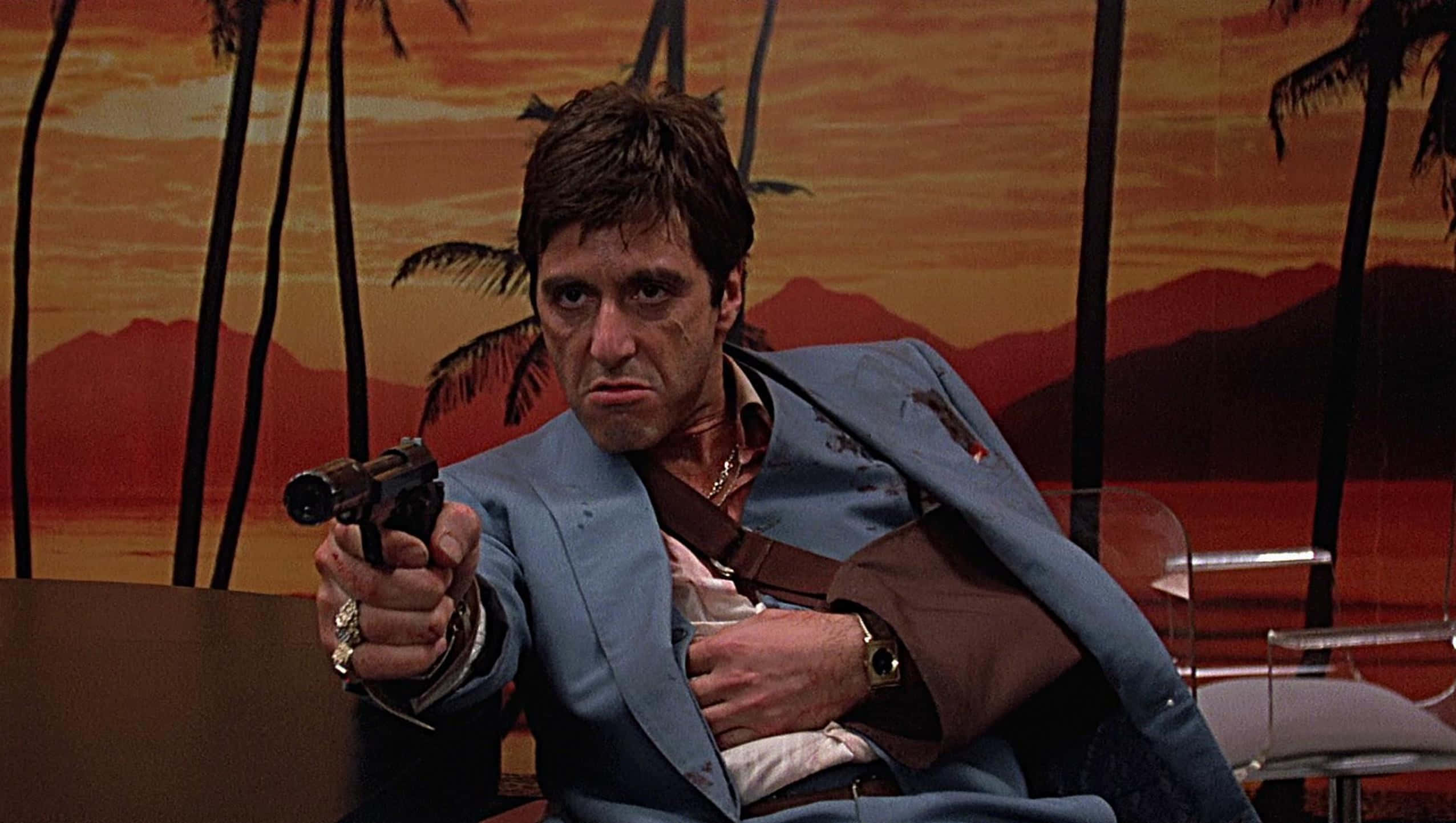 tony montana 桌布 壁紙 — 一個身穿染血藍色西裝的男人，在繪有熱帶日落背景前，以堅定的眼神舉槍瞄準。